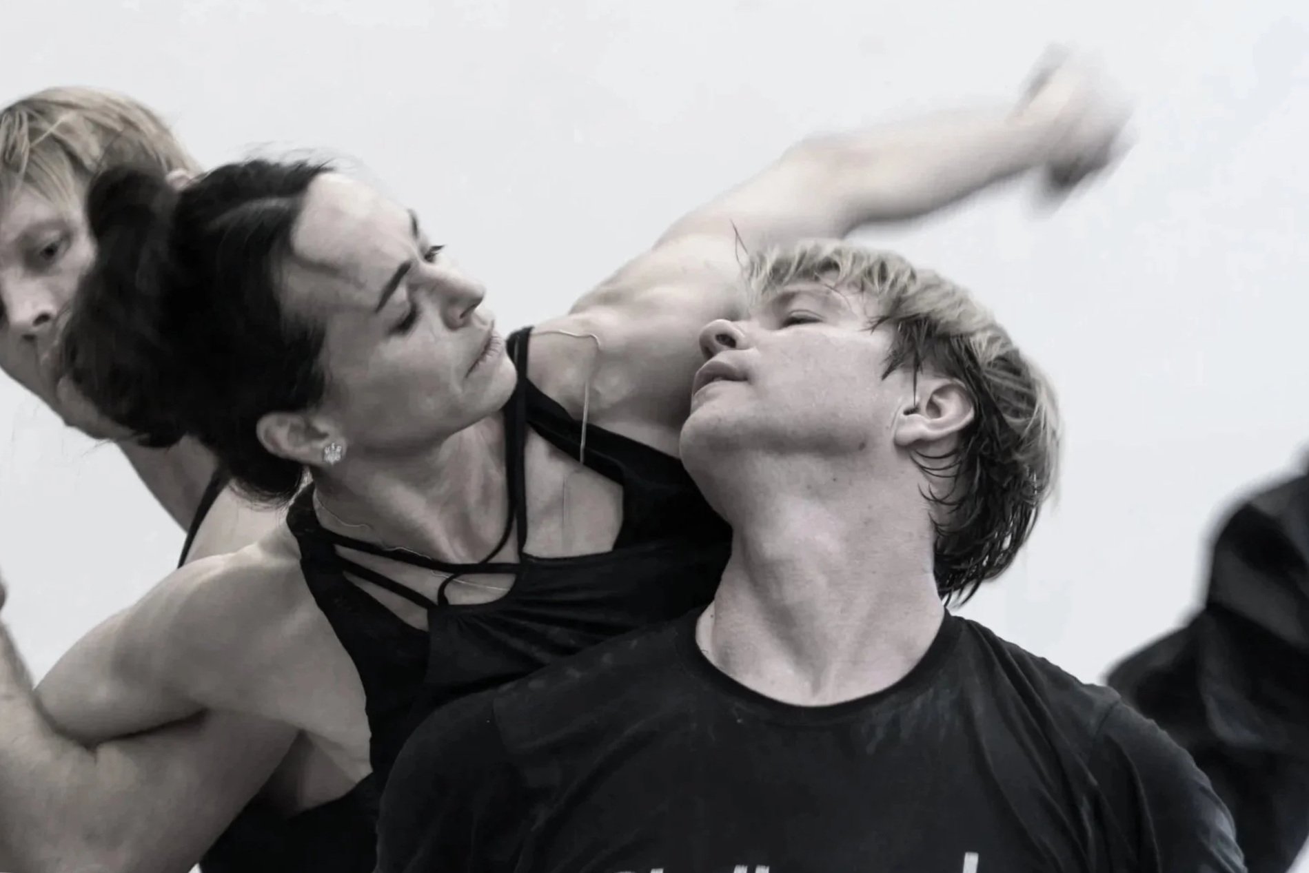 A woman and a man engaged in a dance pose, with the woman leaning over the man and the man tilting his head back, both appearing concentrated, with other dancers partially visible in the background.
