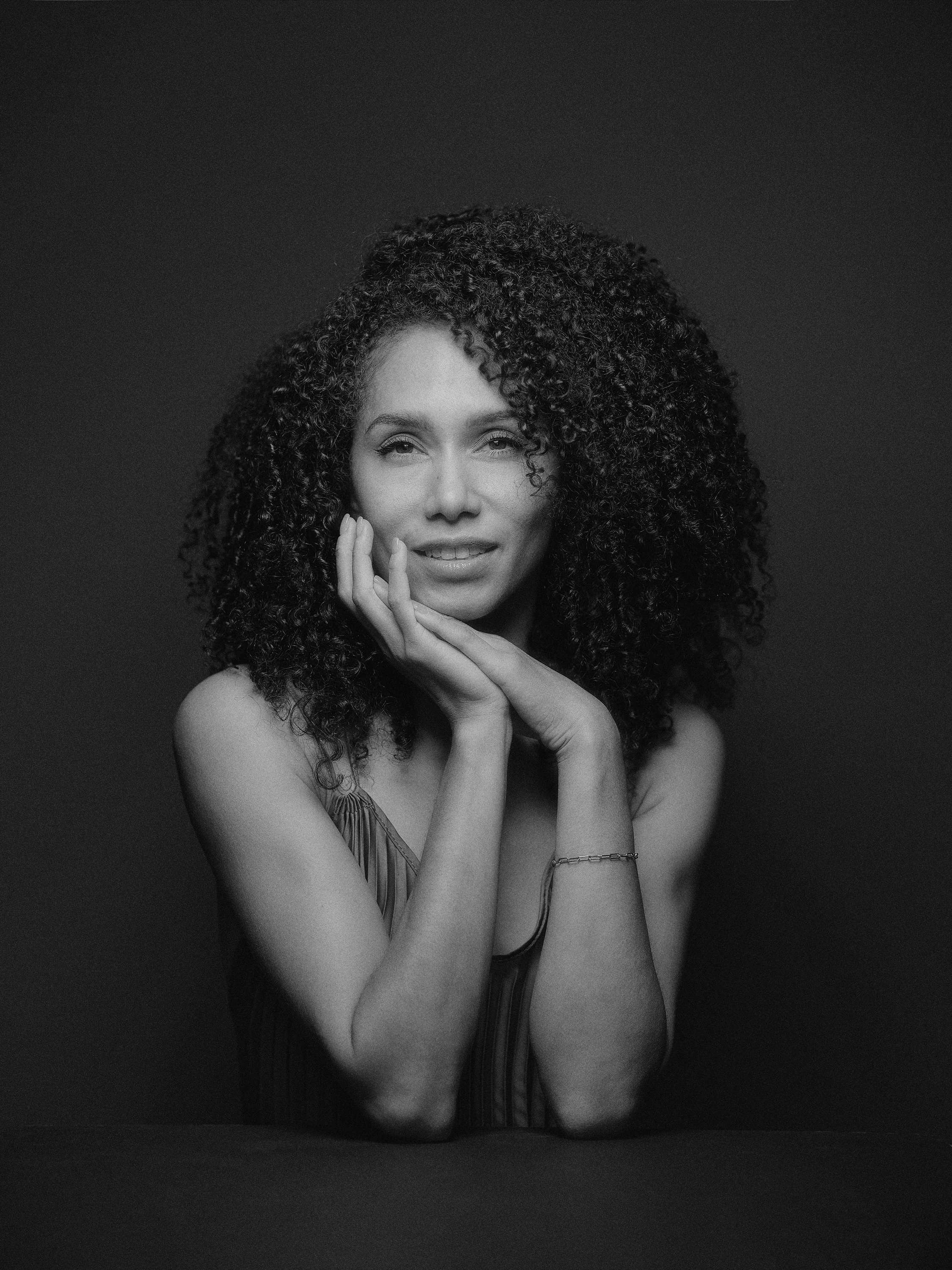 A woman with curly hair, resting her chin on her hand, looking at the camera, in black and white.