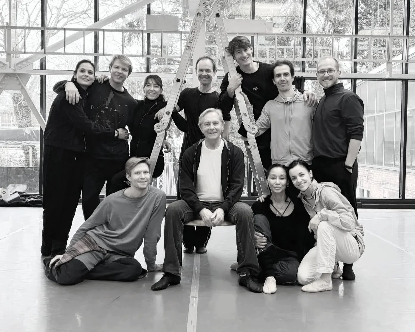 Group of eleven people posing inside a gymnasium with ladders, large windows, and trees outside in the background.