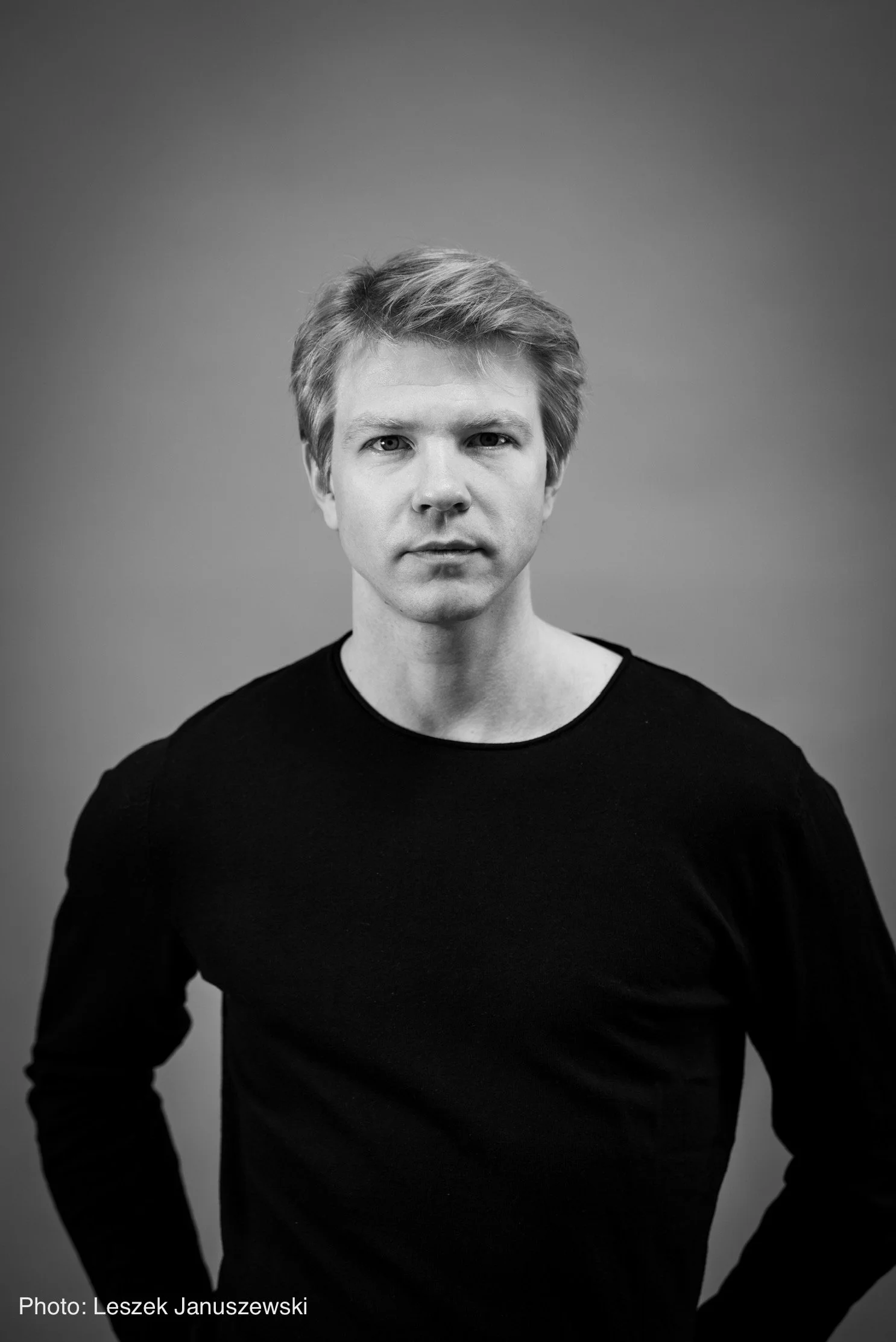 Black and white portrait of a young man with light hair, wearing a black shirt, looking directly at the camera with a neutral expression, against a plain background.