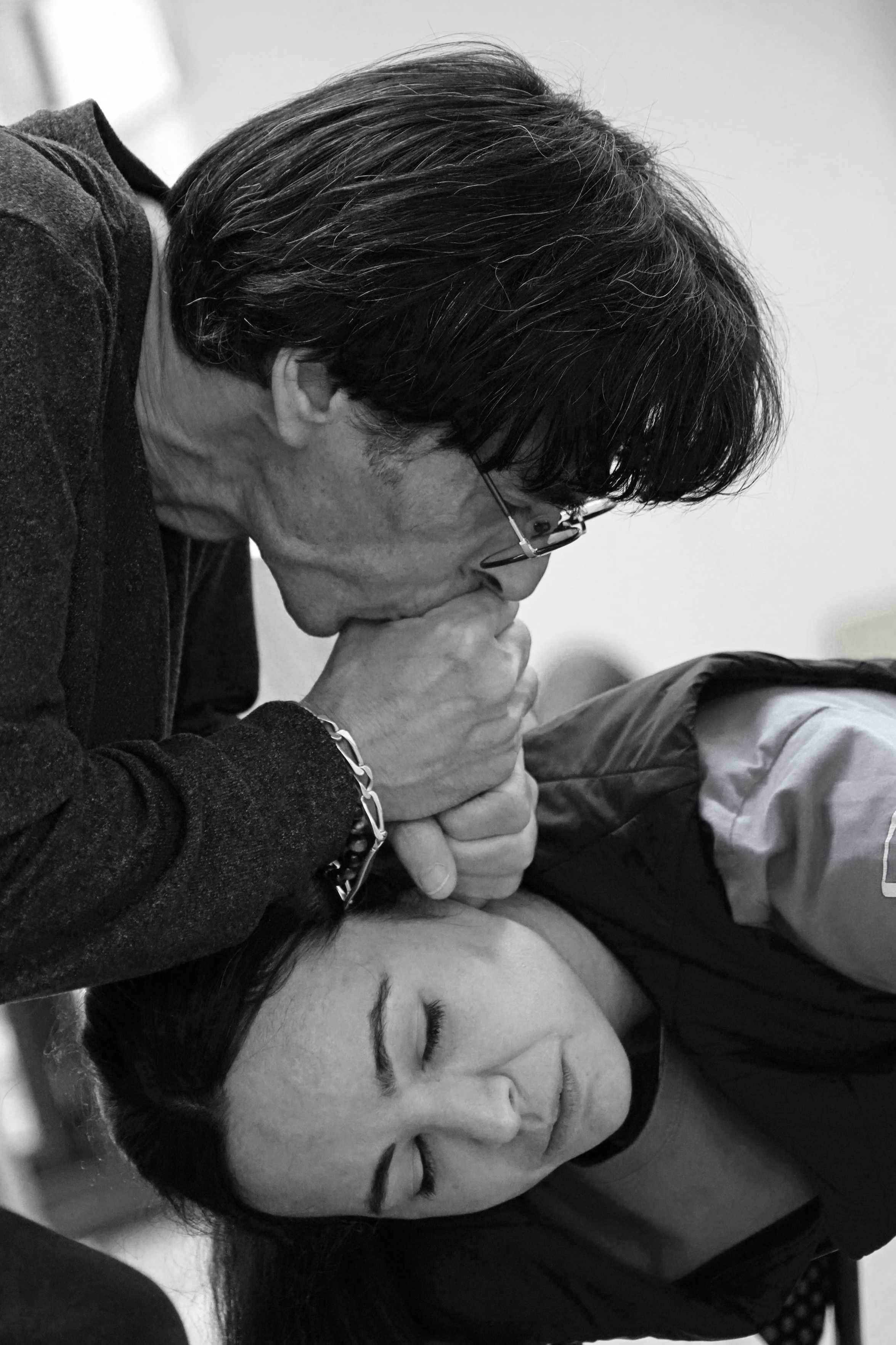 A black and white photo of a man with glasses who appears to be praying or in deep thought, with his hands clasped near his face, leaning over a woman whose eyes are closed, possibly resting or sleeping.
