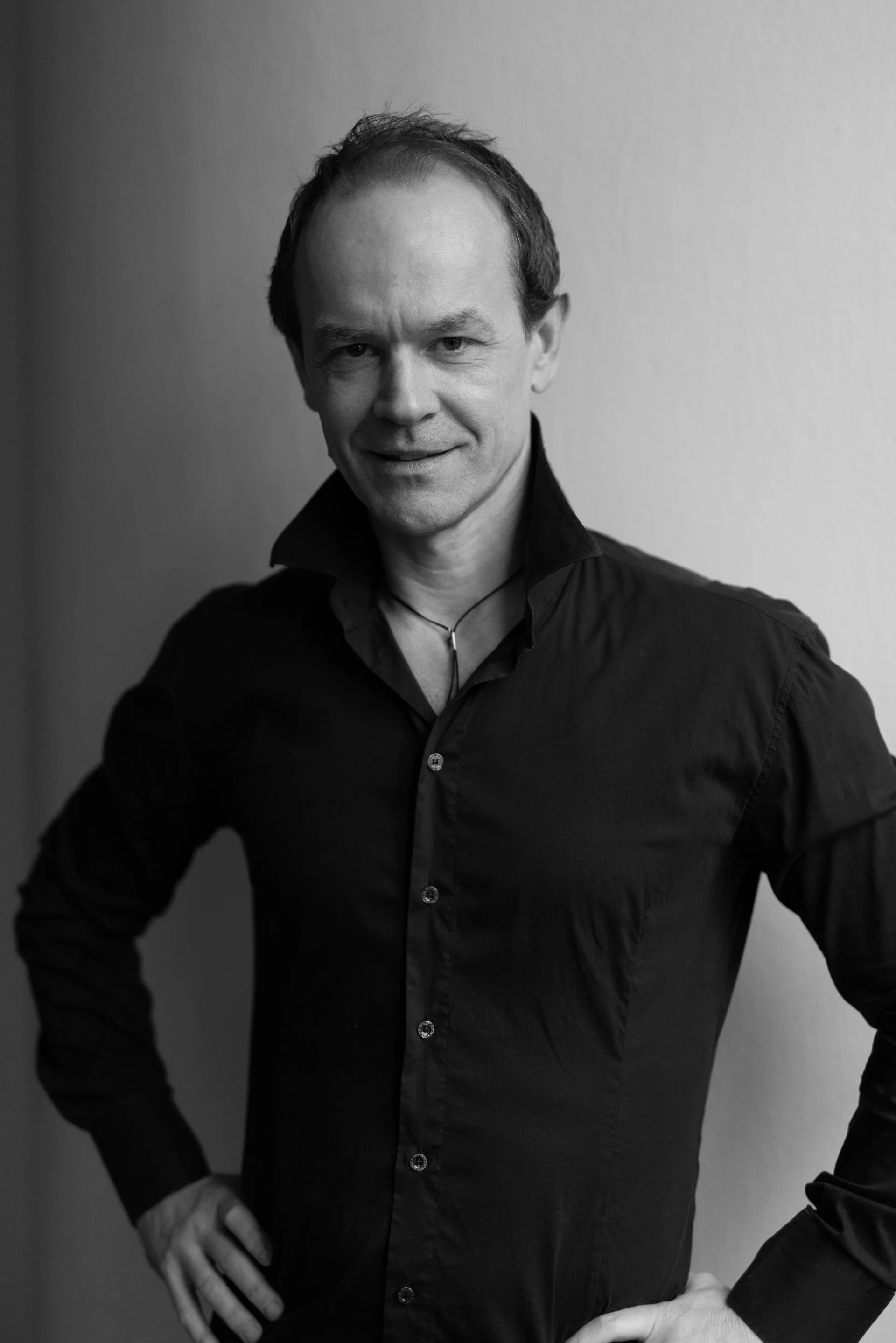 Black and white portrait of a man in a black shirt standing against a plain wall, smiling slightly with his hands on his hips.