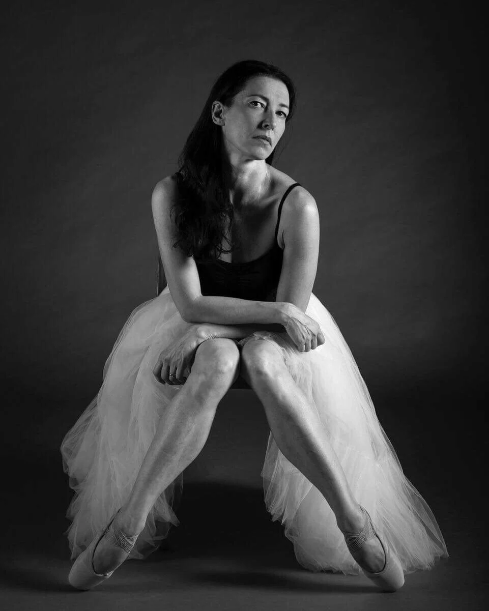A black and white portrait of a woman sitting with her legs crossed and arms resting on her knees, wearing a ballet tutu and pointe shoes, looking thoughtfully at the camera.