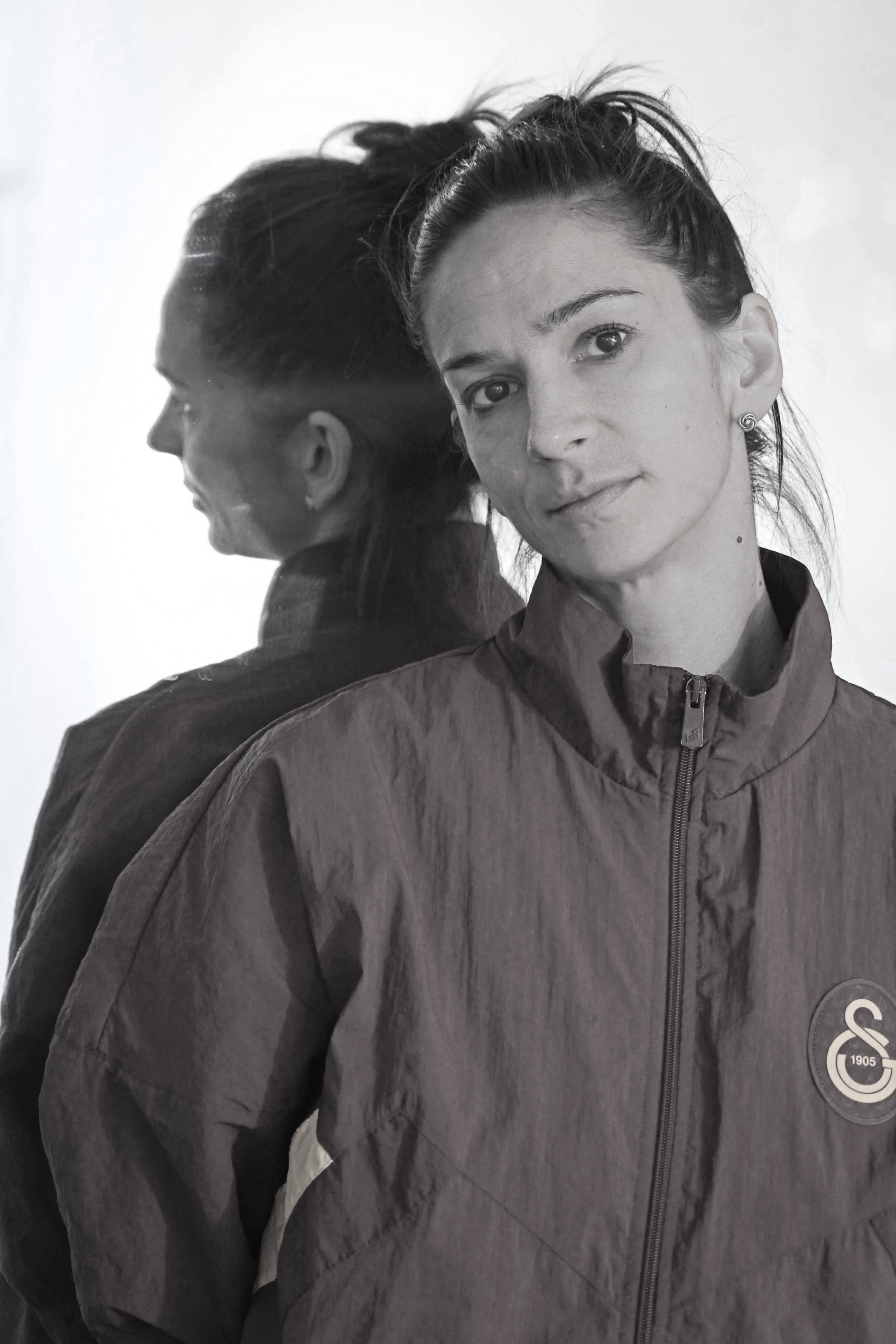 Black and white photo of a woman with her reflection visible in a mirror or glass, wearing a sports jacket with a logo on it.