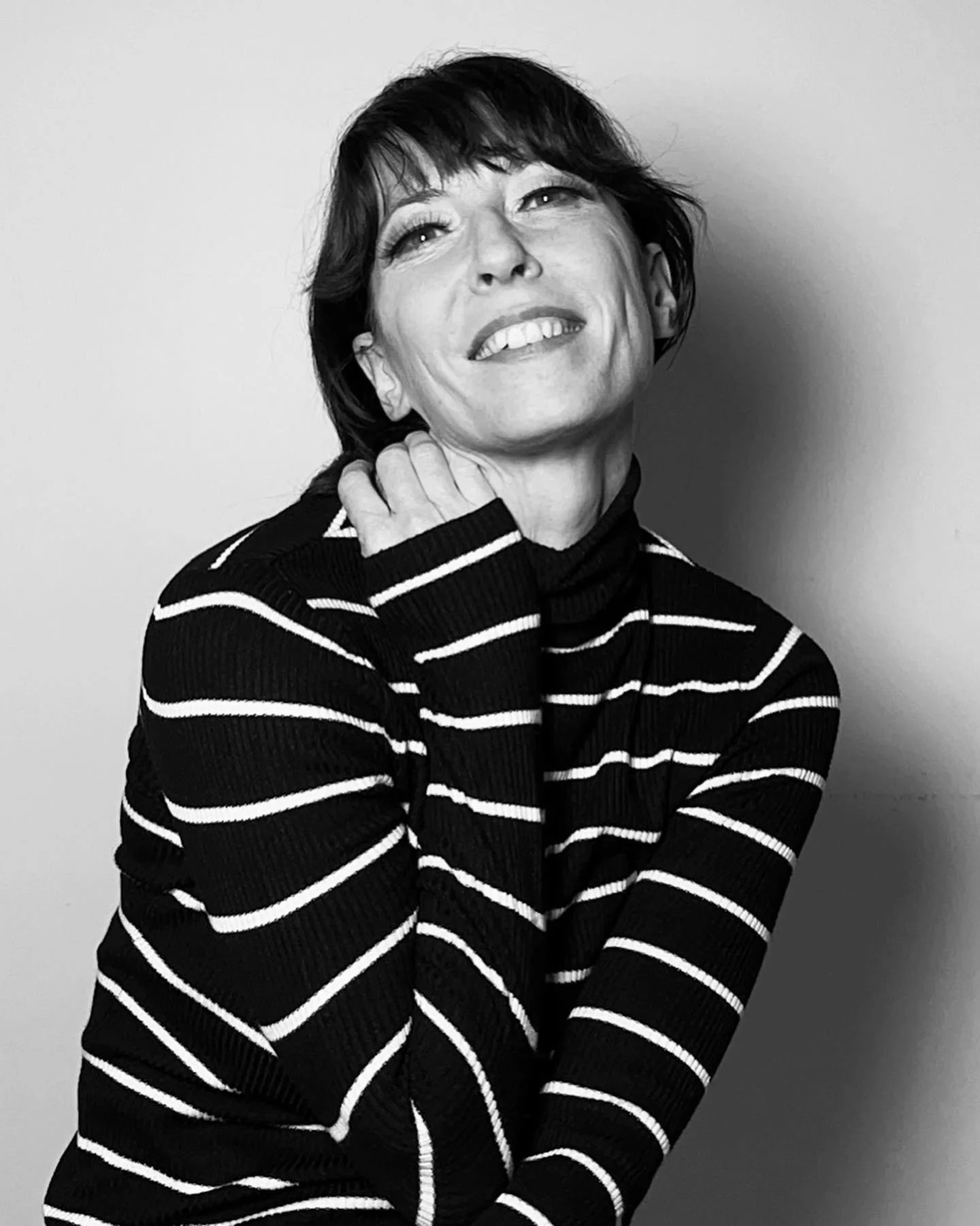 Black and white photo of a woman with short dark hair, smiling, wearing a striped turtleneck sweater, with her hand touching her neck.