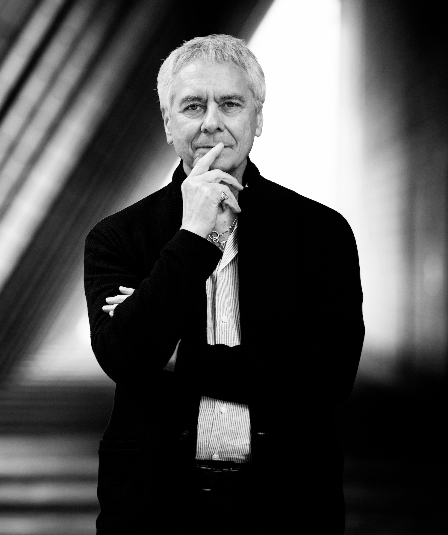 A black and white portrait of an older man with short, light-colored hair, wearing a dark jacket and light shirt, standing outdoors with an abstract, curved architectural background. He is thoughtfully touching his chin with his right hand.