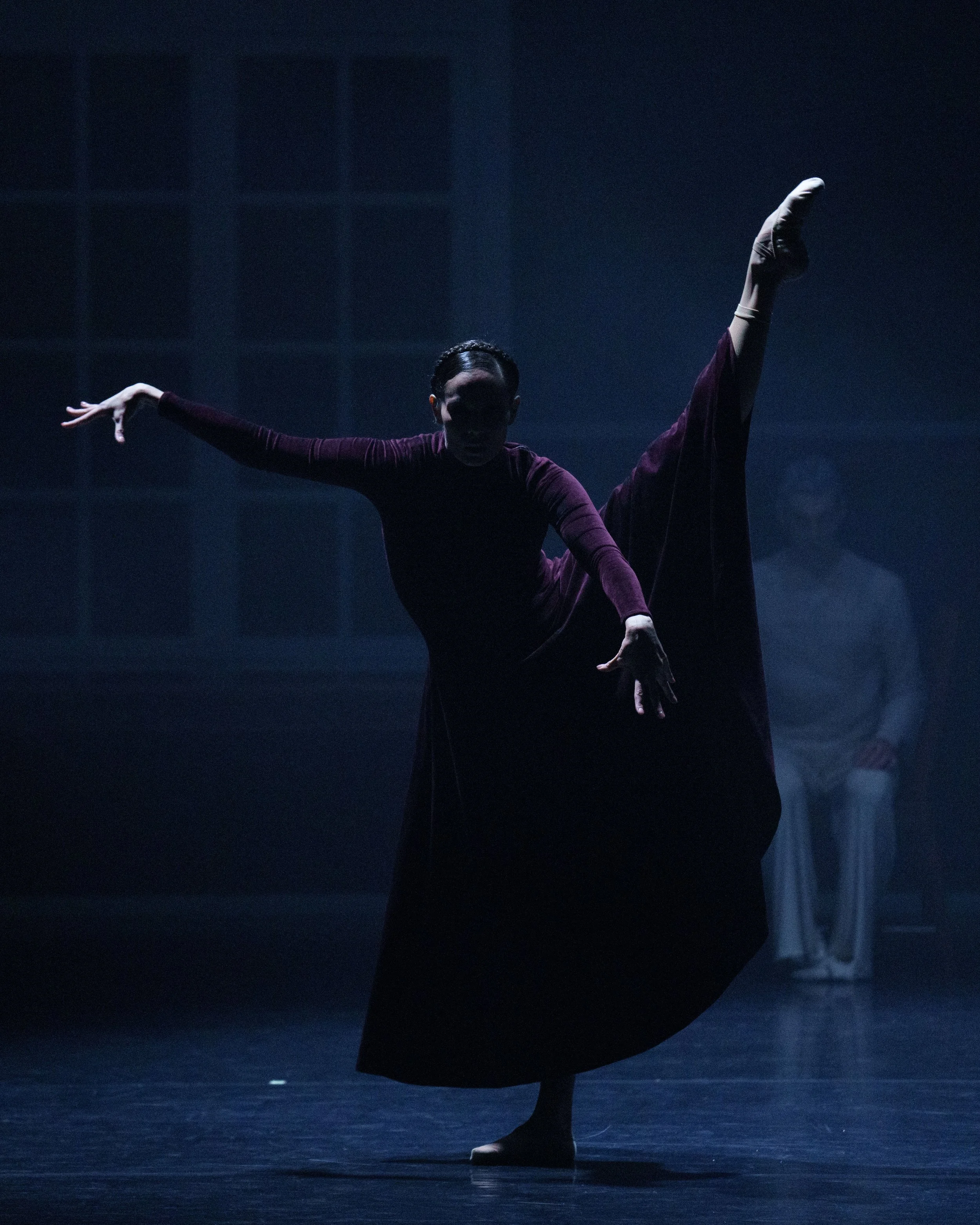 A dancer performing on stage with dramatic lighting, wearing a long dark dress with one leg raised high and extended sideways.