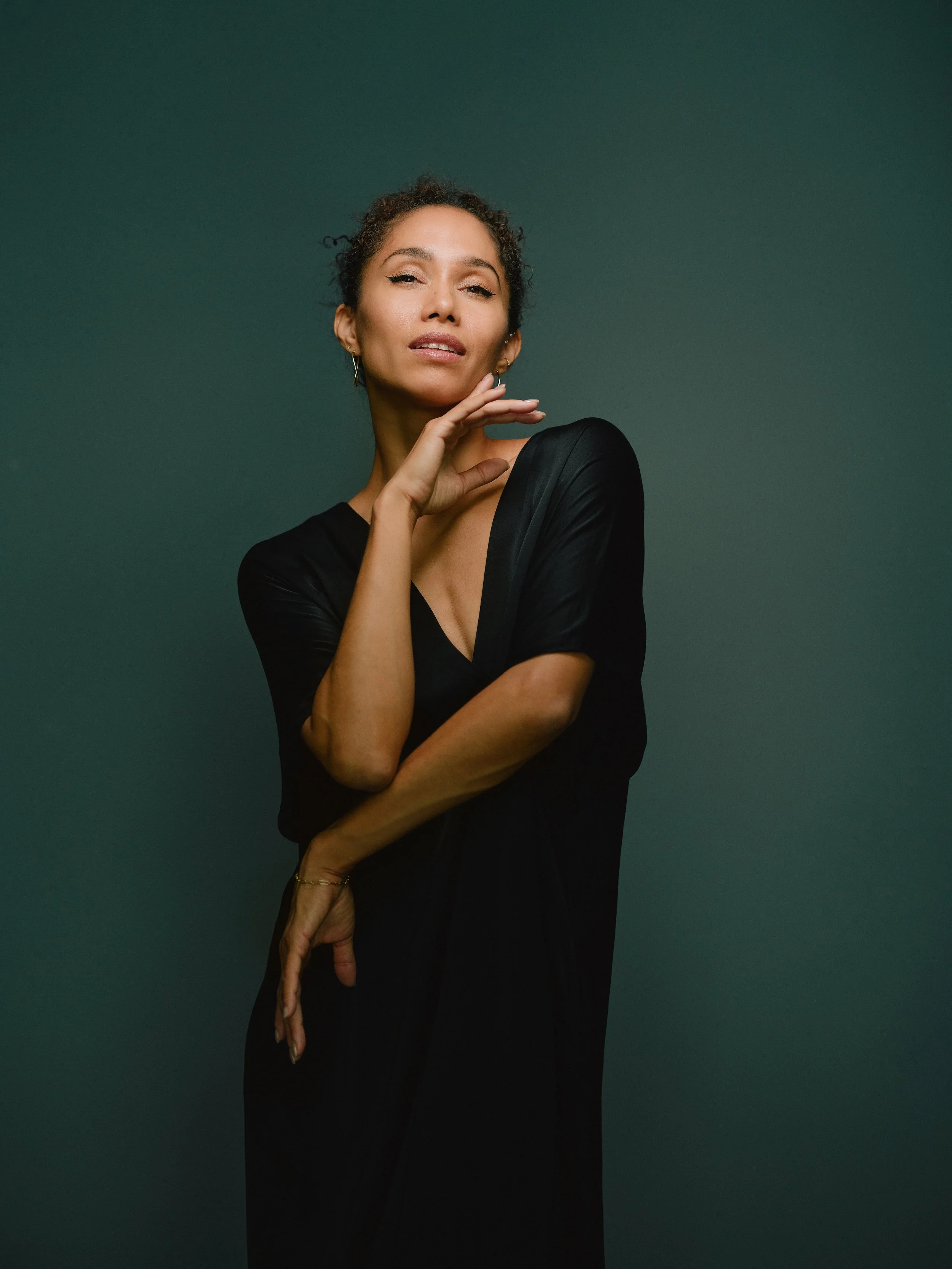 Portrait of a woman with short curly hair, wearing a black dress, posing against a green background.
