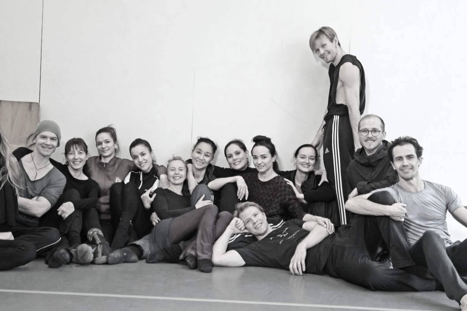 Group of smiling people in athletic wear sitting and lying on the floor, with one person standing on a ledge, in an indoor workout space.