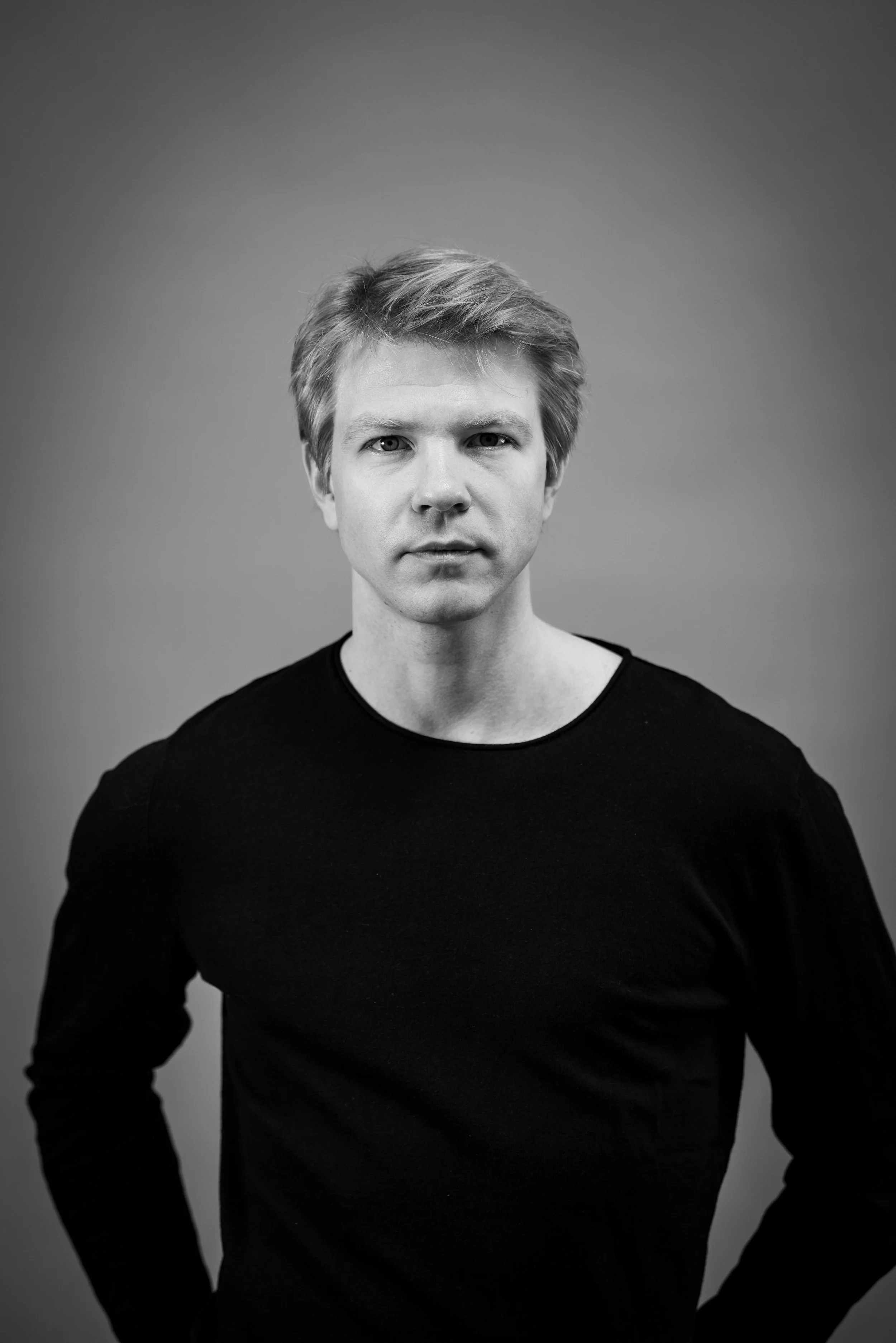 Black and white portrait of a young man with light hair, wearing a black long-sleeve shirt, standing against a plain background.