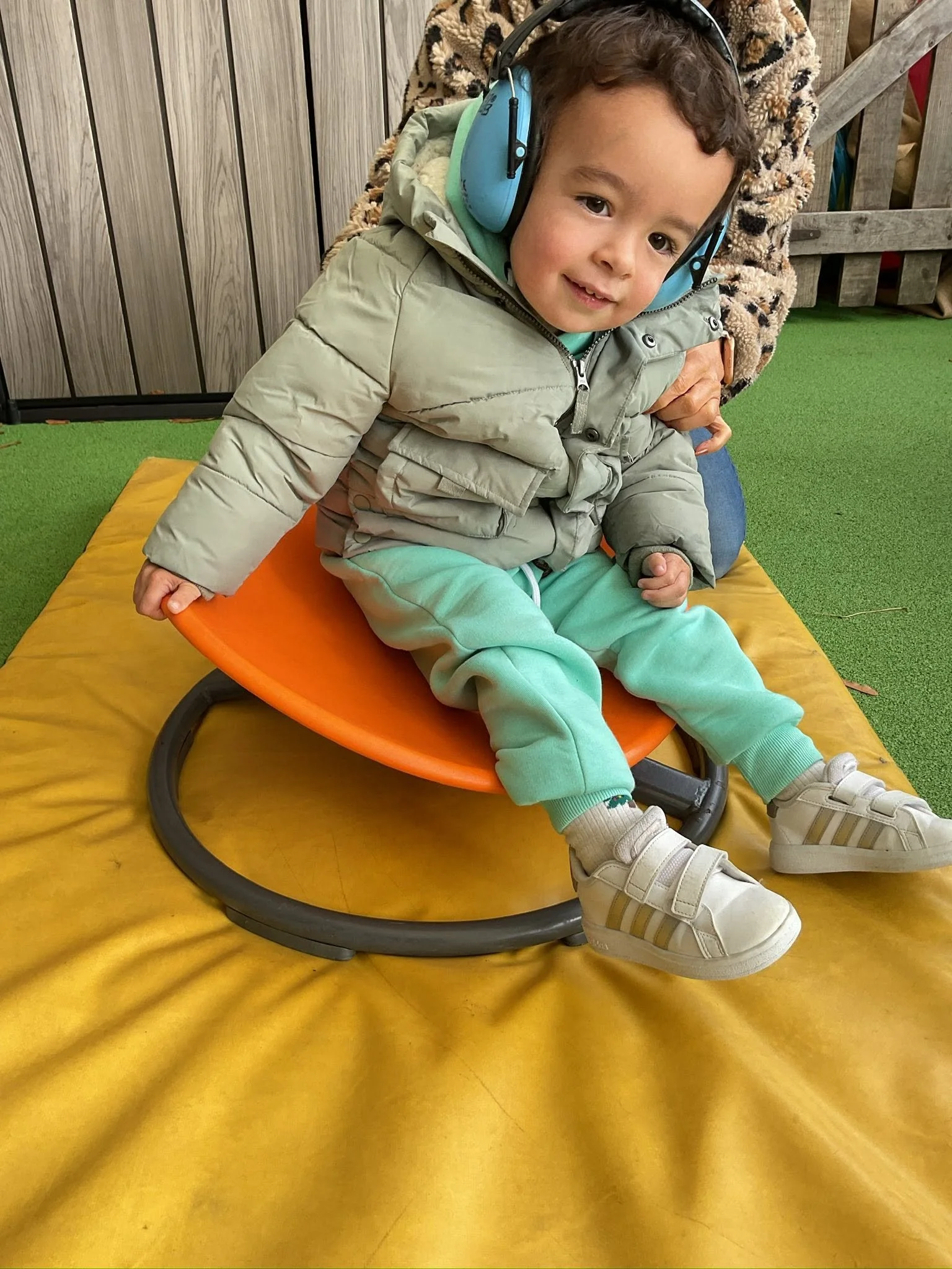 A young boy in a light green hoodie and matching sweatpants, wearing a beige puffer jacket and white sneakers, is sitting on an orange round bouncy chair on a yellow mat. He's wearing large blue noise-canceling headphones, smiling, with a wooden fence and green artificial grass in the background.