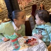 A woman and a young boy are sitting at a table, facing each other. The woman has her hair tied back and is smiling at the boy. The boy is wearing a light blue hoodie with the Moschino logo. There is a plate of assorted fruits and a turquoise water bottle on the table. In the background, other people are seated and talking in a restaurant or café setting.