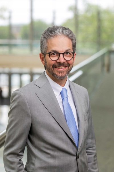 Smiling man in a gray suit and blue tie standing indoors with glass railing and blurred background.