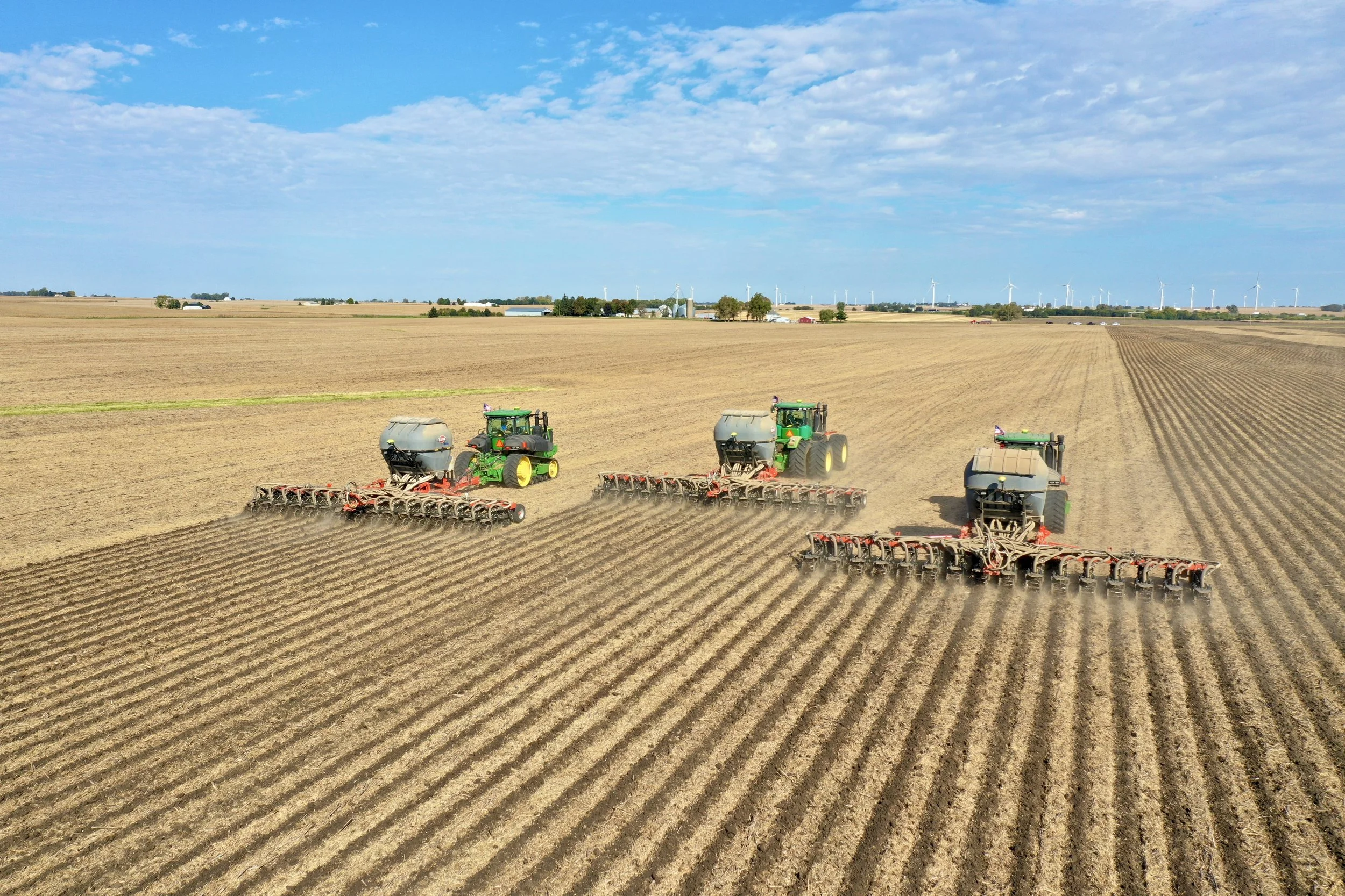 A large green John Deere tractor plowing a field with a red and white seed planter attached, under a blue sky with scattered clouds.