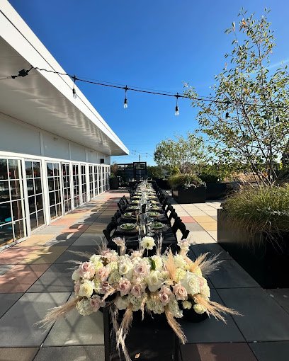 Outdoor patio with a long dining table decorated with flowers, string lights overhead, and surrounding greenery under a clear blue sky.
