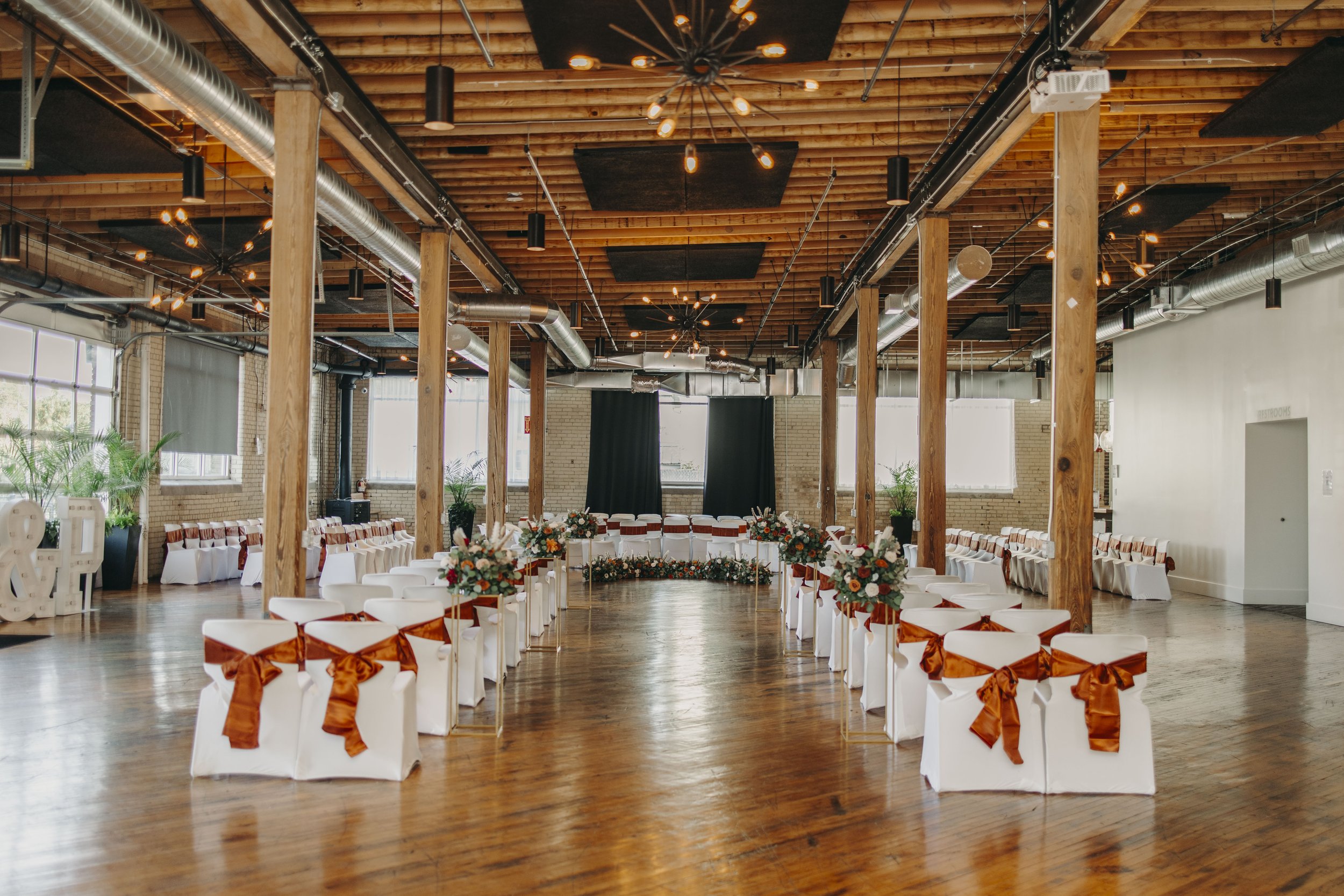 Spacious event venue with wooden beams and polished floors, set up for a wedding with white chairs adorned with brown bows, floral arrangements, and ambient lighting.