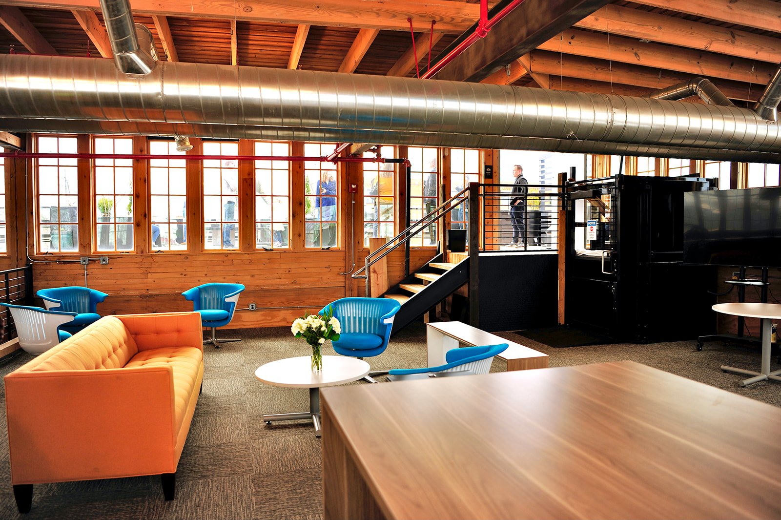 Modern office space with orange sofa, blue chairs, wooden table, and large windows.