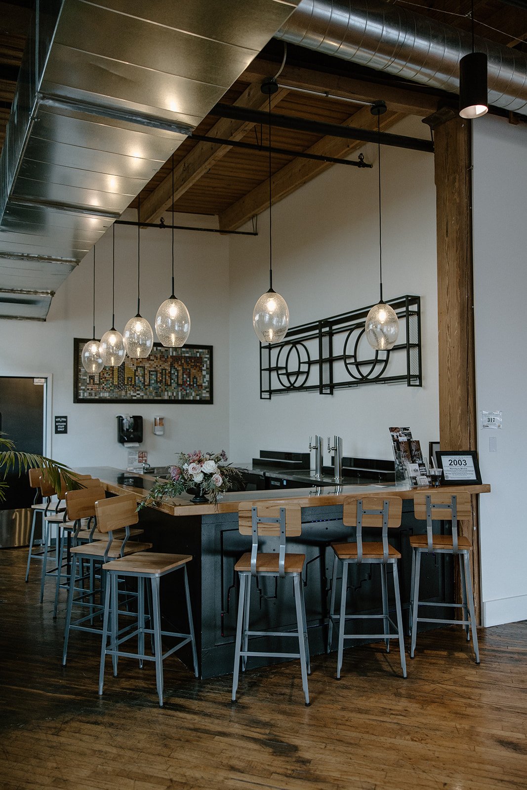 Modern cafe interior with bar, wooden stools, pendant lights, and decorative art on the wall.