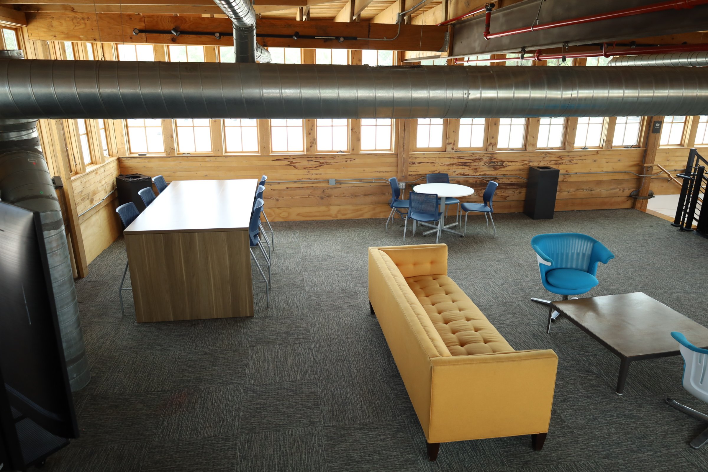 Modern office lounge area with yellow couch, blue chairs, wooden table, and industrial ducting overhead.