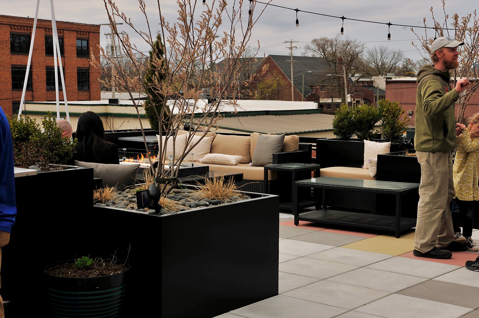 Rooftop patio with seating and plants, featuring a man and child, surrounded by buildings and string lights overhead.