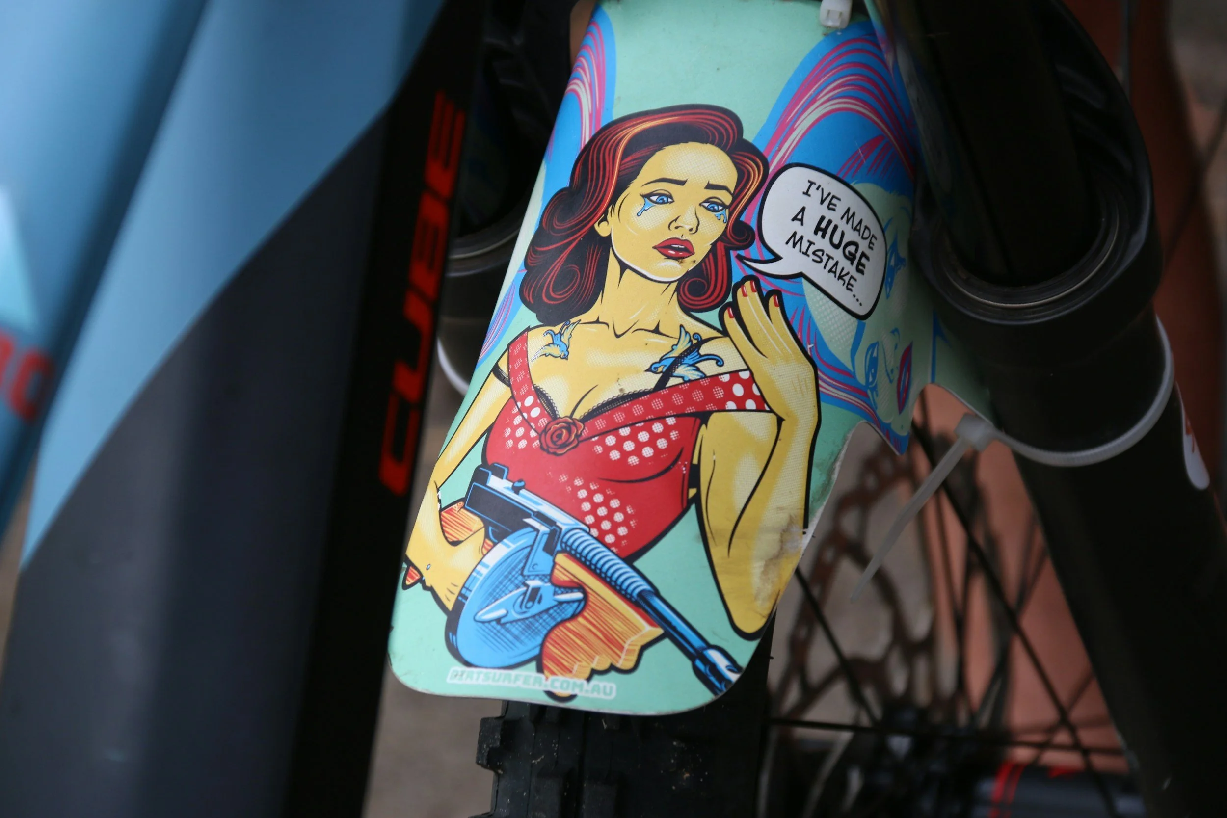 A sticker on a bicycle features a comic-style illustration of a woman with red hair, holding a gun, with tears on her face. She is wearing a red and white polka dot dress with a rose detail and has a speech bubble that says, "I've made a huge mistake."