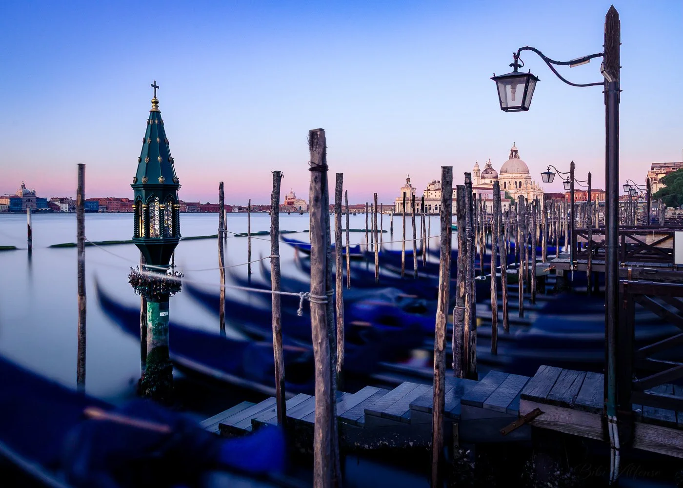 Long exposure shot of gondolas docked at a wooden pier in Venice after sunrise, with Santa Maria della Salute and historic buildings in the background.