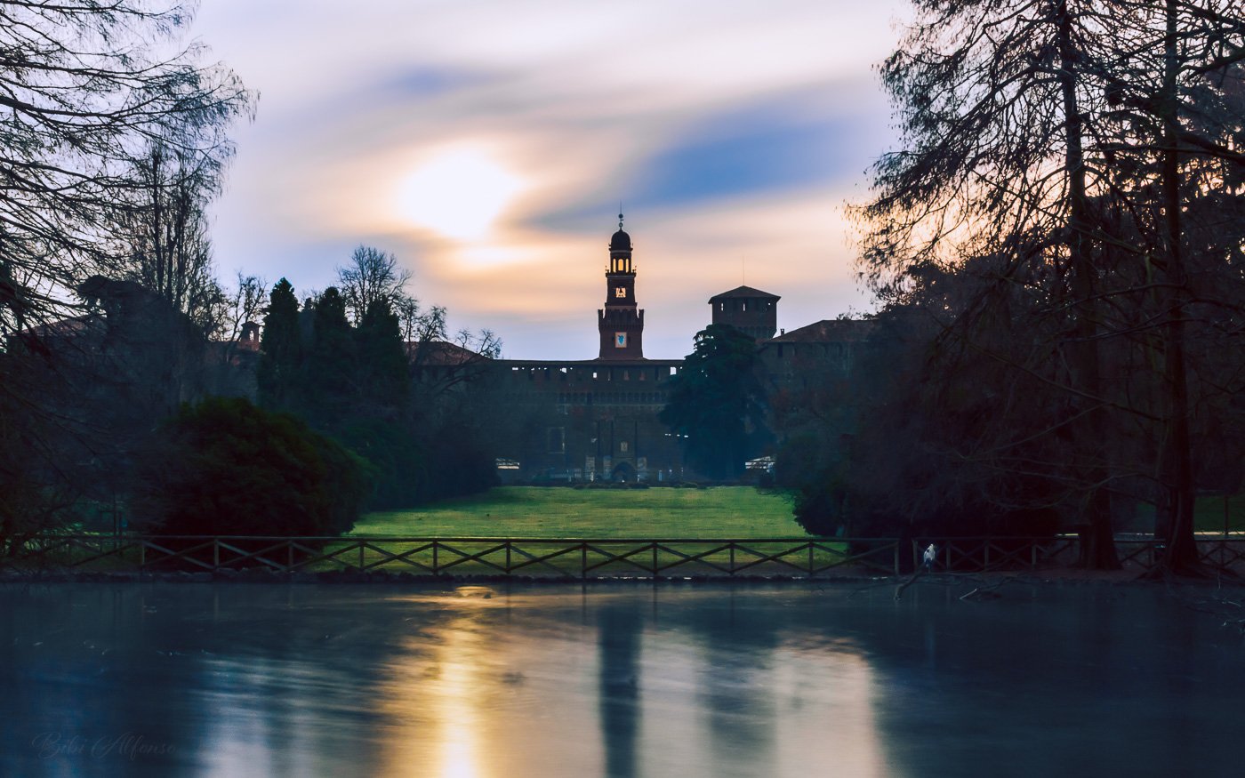 Mist rising over Parco Sempione in Milan during a quiet morning, with lush greens and soft blue tones creating a serene atmosphere.
