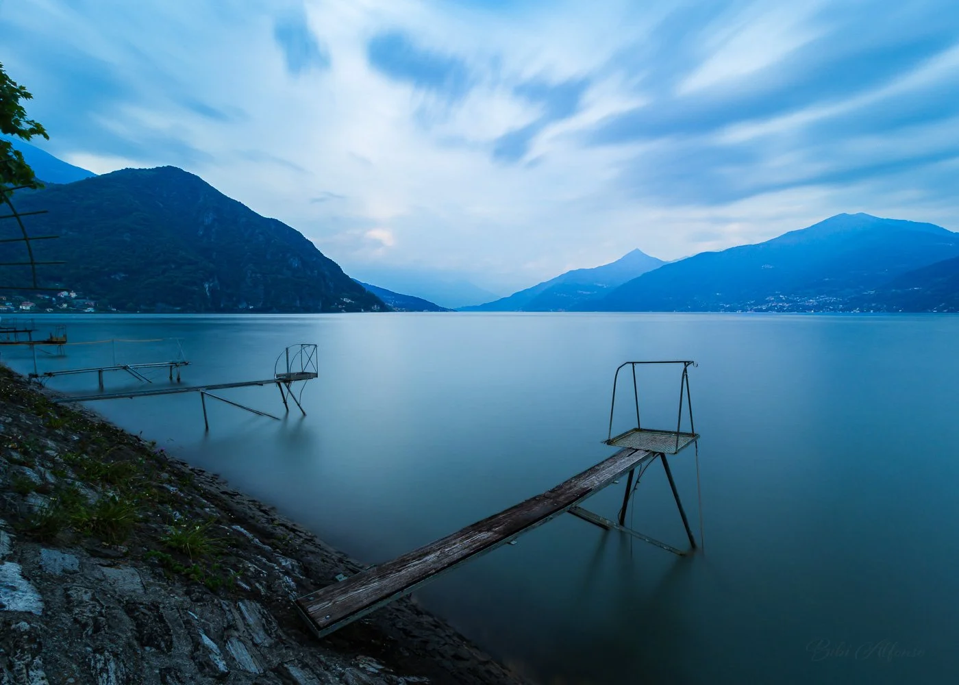 Long exposure of Lake Como at Menaggio, with silky still waters contrasting against drifting clouds in the blue-toned sky, creating a serene yet dynamic atmosphere.
