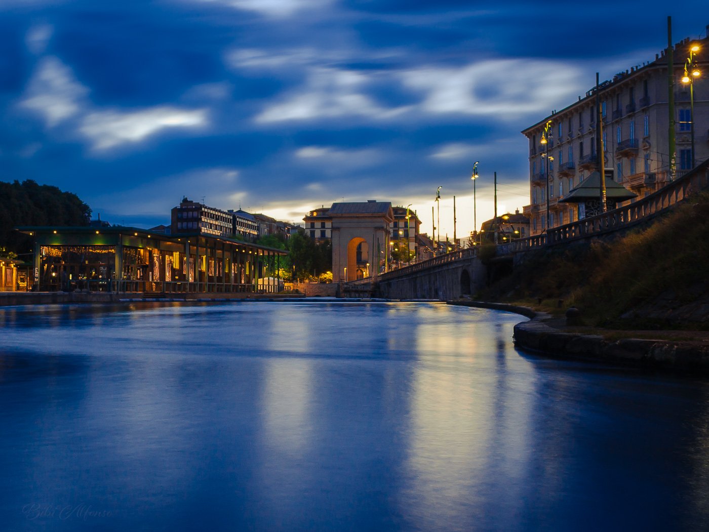 Long exposure at Darsena, Milan, transforming a windy morning into a serene scene, with calm waters reflecting golden lights from surrounding buildings under deep blue skies.
