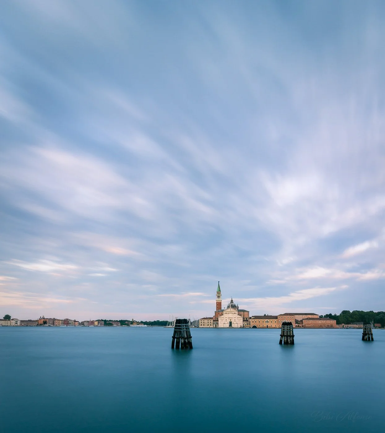 Long exposure golden hour view of San Giorgio Maggiore in Venice, Italy, with smooth water, softly moving clouds, and warm sunlight illuminating the historic church and waterfront buildings in a serene travel photography scene.