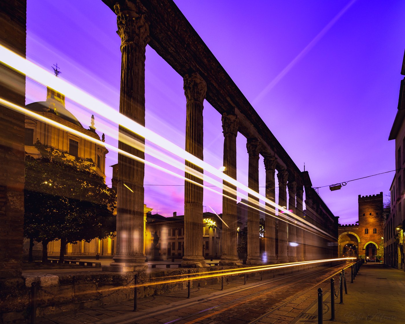 Very early morning long exposure at Colonne di San Lorenzo in Milan, with golden tram light trails weaving past the ancient Roman columns under a blue and purple sky.