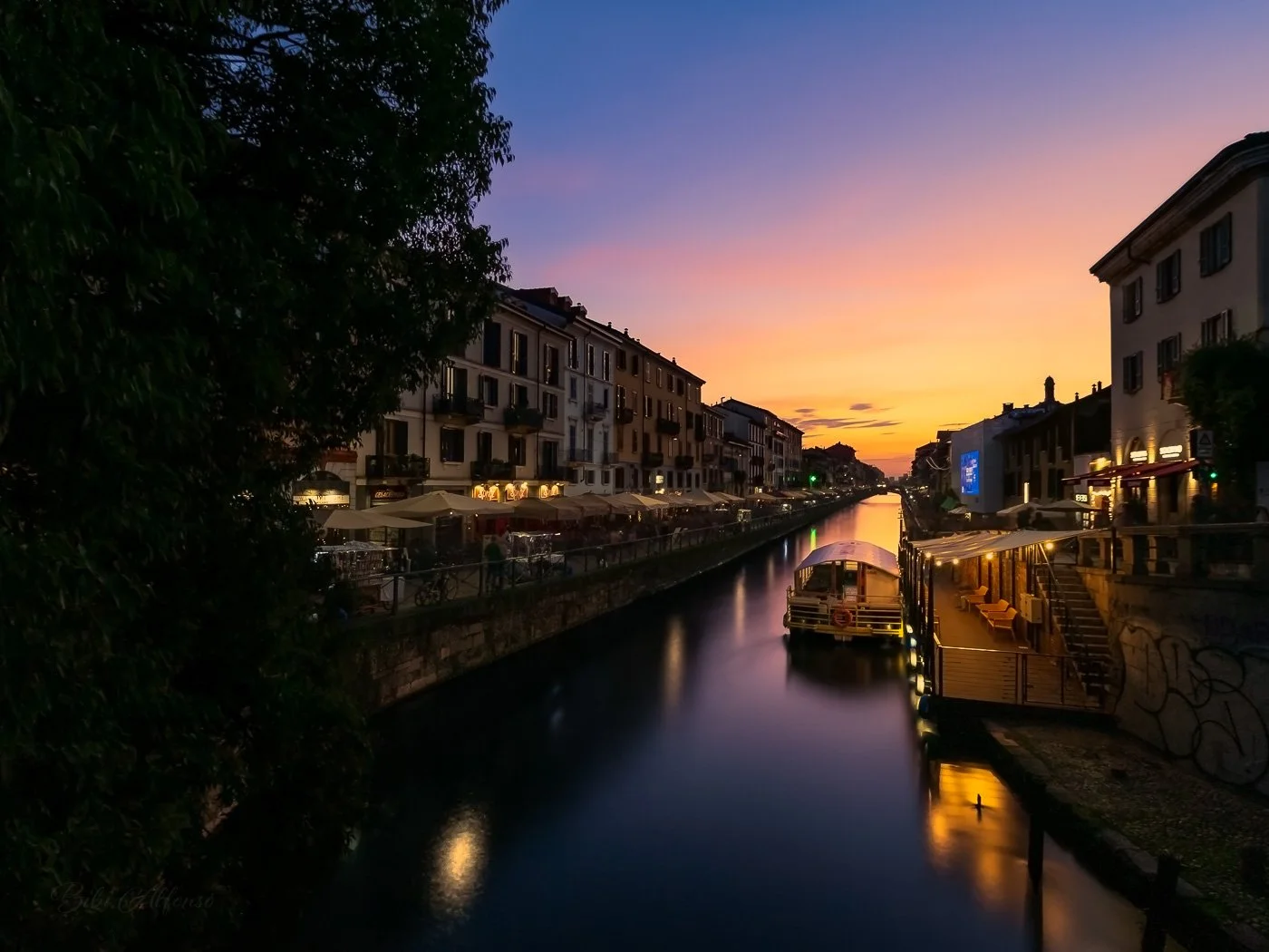 Two minutes long exposure twilight shot of Naviglio Grande canal in Milan, Italy, showing colorful buildings, outdoor cafes with umbrellas, boats, and streetlights glowing under sunset skies.