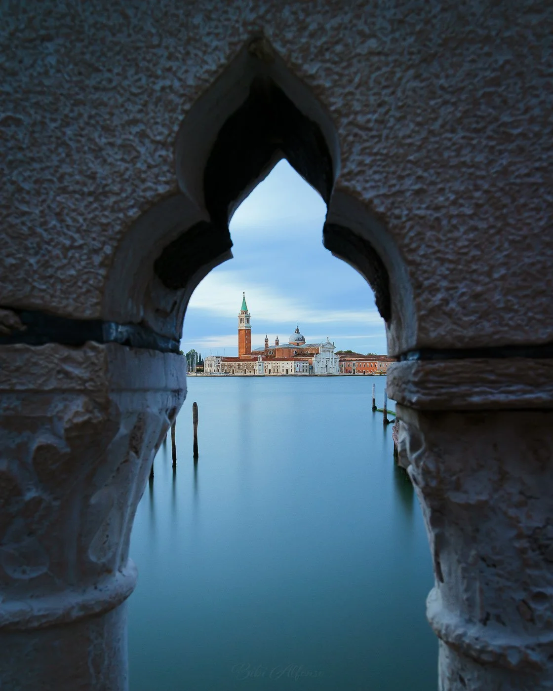 Long exposure view of San Giorgio Maggiore in Venice, Italy, framed through the ornate stone arch of Ponte della Paglia, with smooth water, soft blue tones, and historic architecture illuminated under a calm sky.