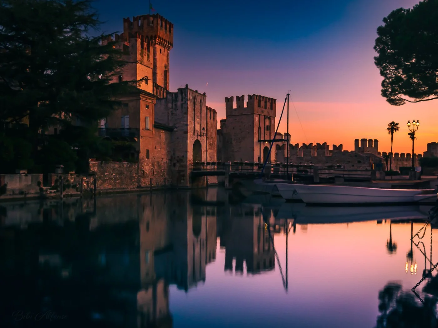 Long exposure of Scaliger Castle in Sirmione at sunrise, with soft orange light illuminating the stone walls against a cool blue morning sky.