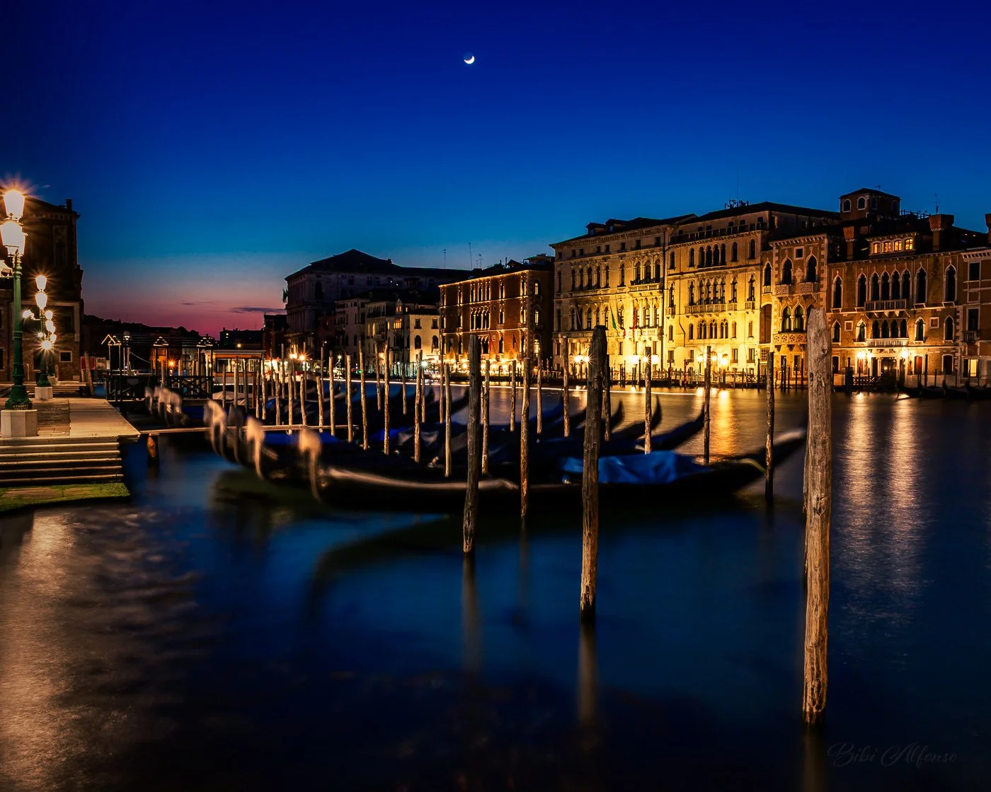 Long exposure after-sunset shot of the Grand Canal from Santa Maria della Salute, Venice, Italy, featuring gondolas docked by wooden posts, illuminated historic buildings, and a crescent moon against a deep blue sky.

