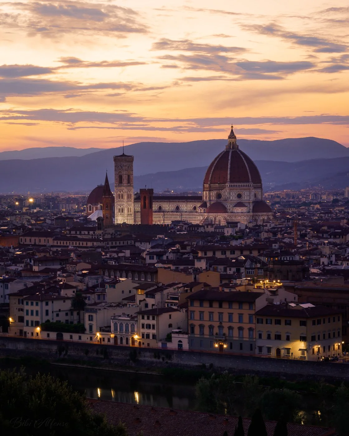 Florence at sunset, showing the Cathedral of Santa Maria del Fiore and Giotto’s Campanile lit above the city, with rooftops, river, and distant mountains under a warm evening sky.