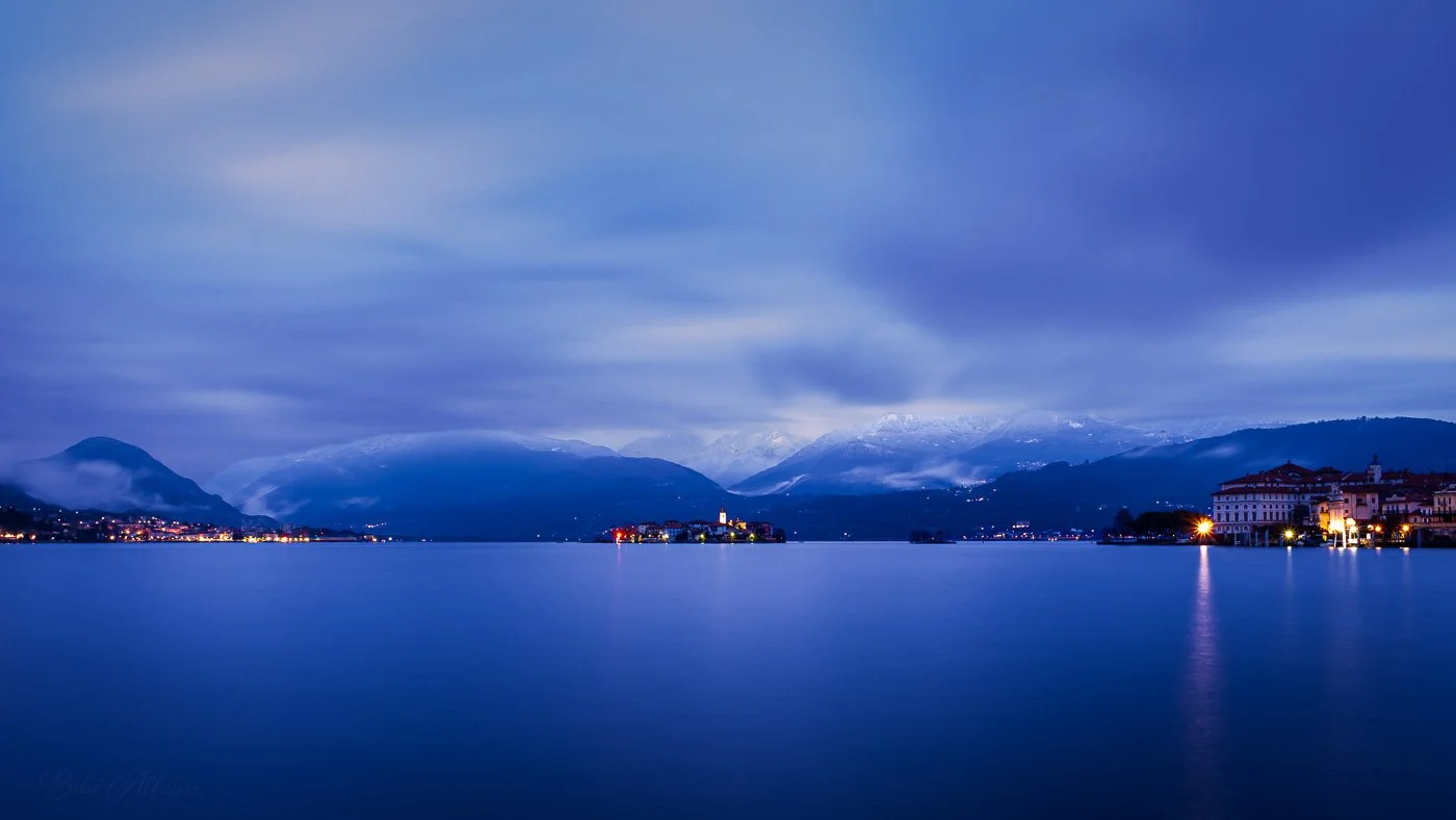 “Blue-hour long exposure before dawn featuring Isola Pescatori and Isola Bella on Lake Maggiore, with a deep blue sky, soft streaky clouds, and gentle artificial lights illuminating the islands.