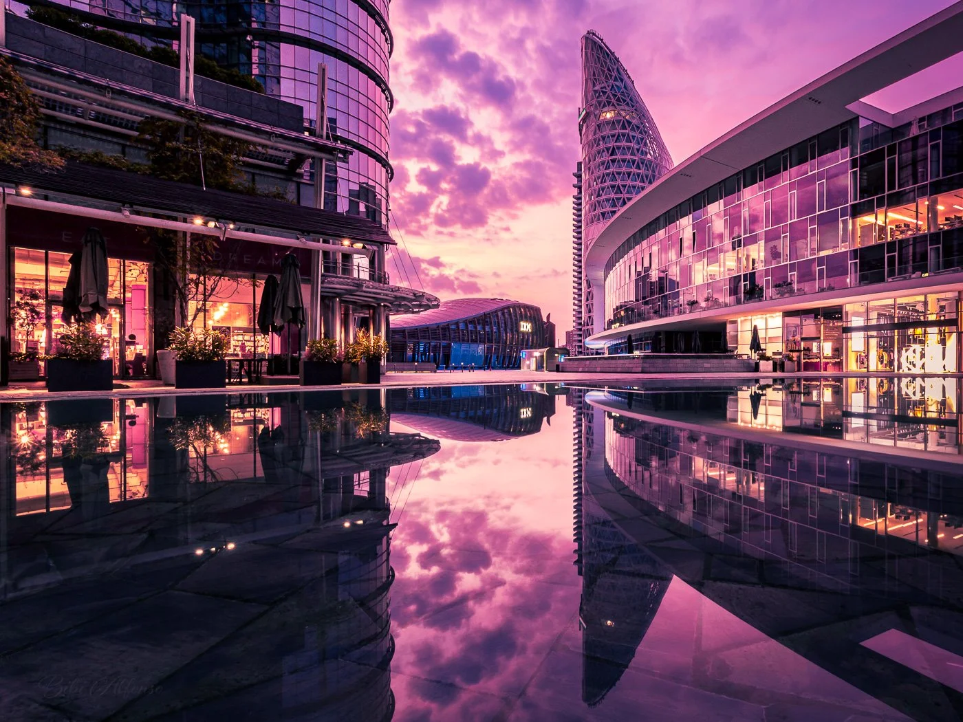 Pink sunrise illuminating Piazza Gae Aulenti in Milan, with reflections of modern architecture against a pink and gold sky.
