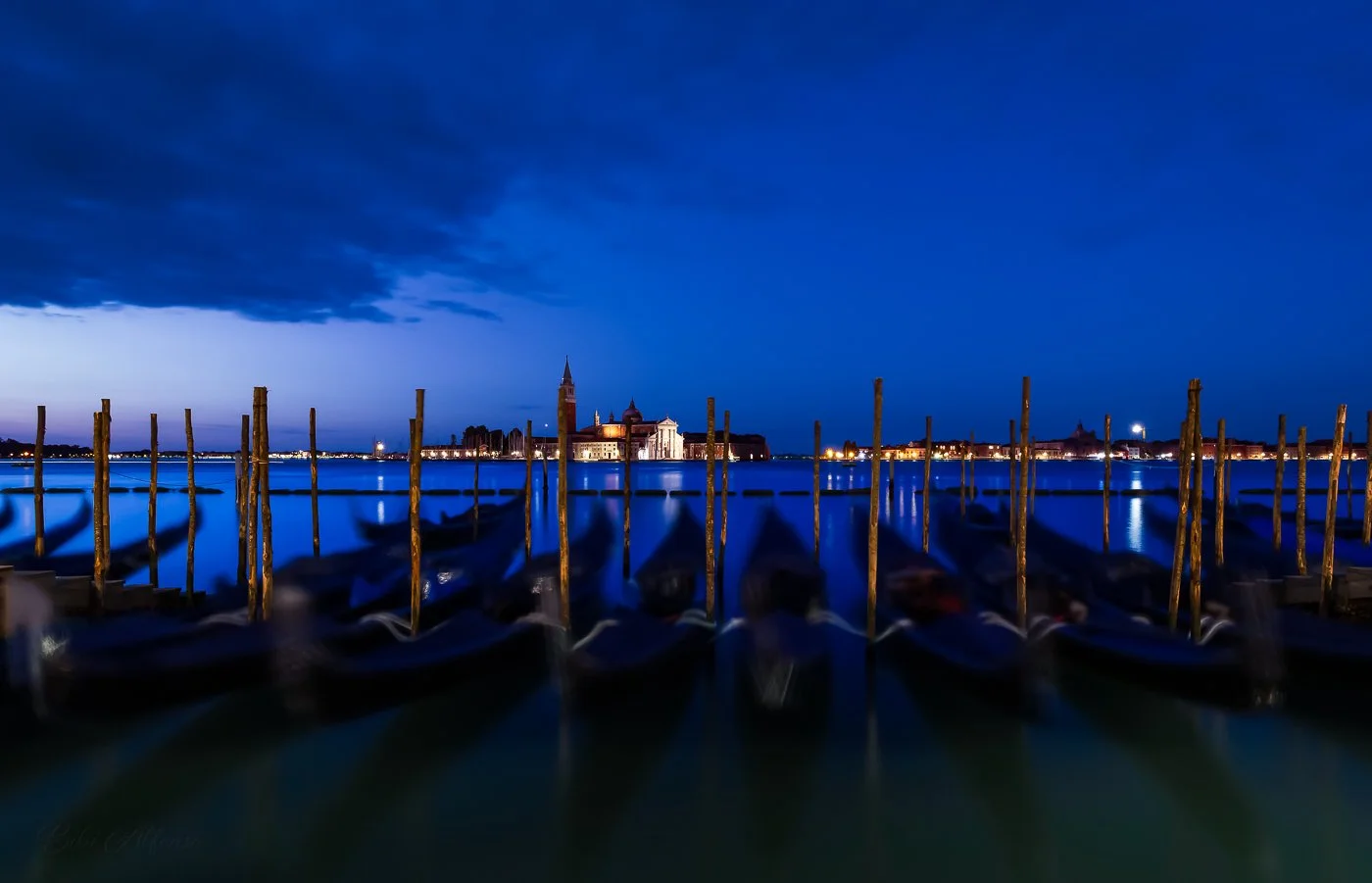 Long-exposure blue-hour photograph of gondolas and wooden mooring poles facing San Giorgio Maggiore across the Venetian lagoon, with deep cobalt sky reflections and soft motion blur in the foreground boats.