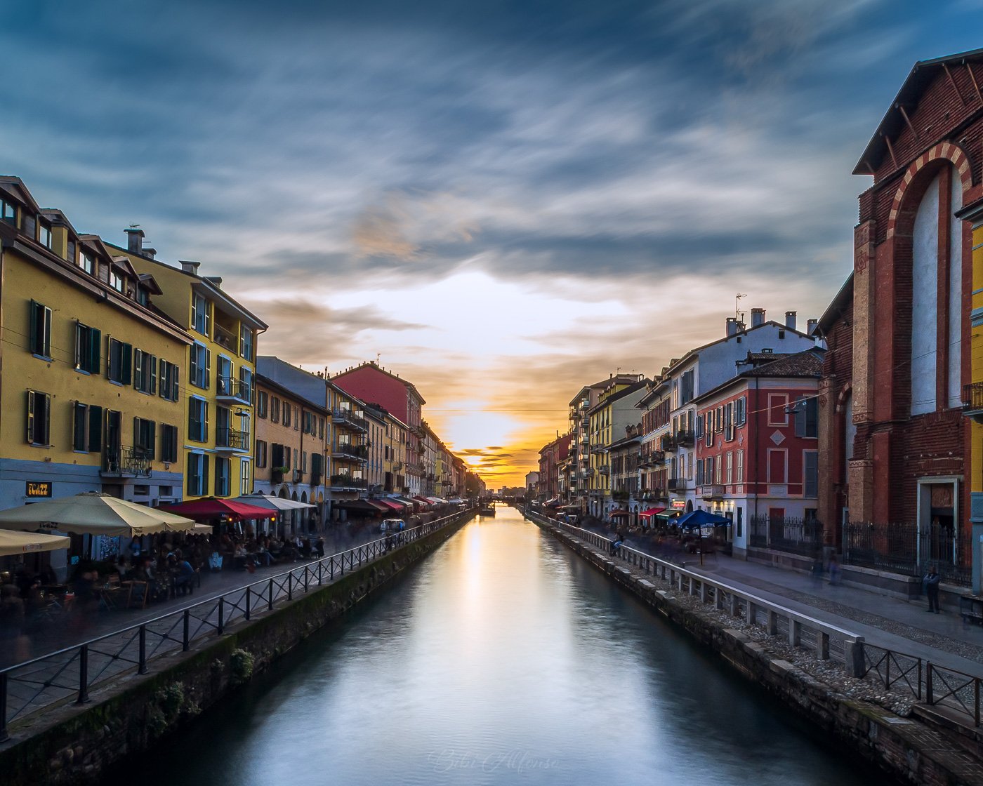 30-second long exposure of a sunset over Naviglio Grande, Milan, featuring silky water reflecting blue and yellow tones, with surrounding red brick buildings adding contrast.