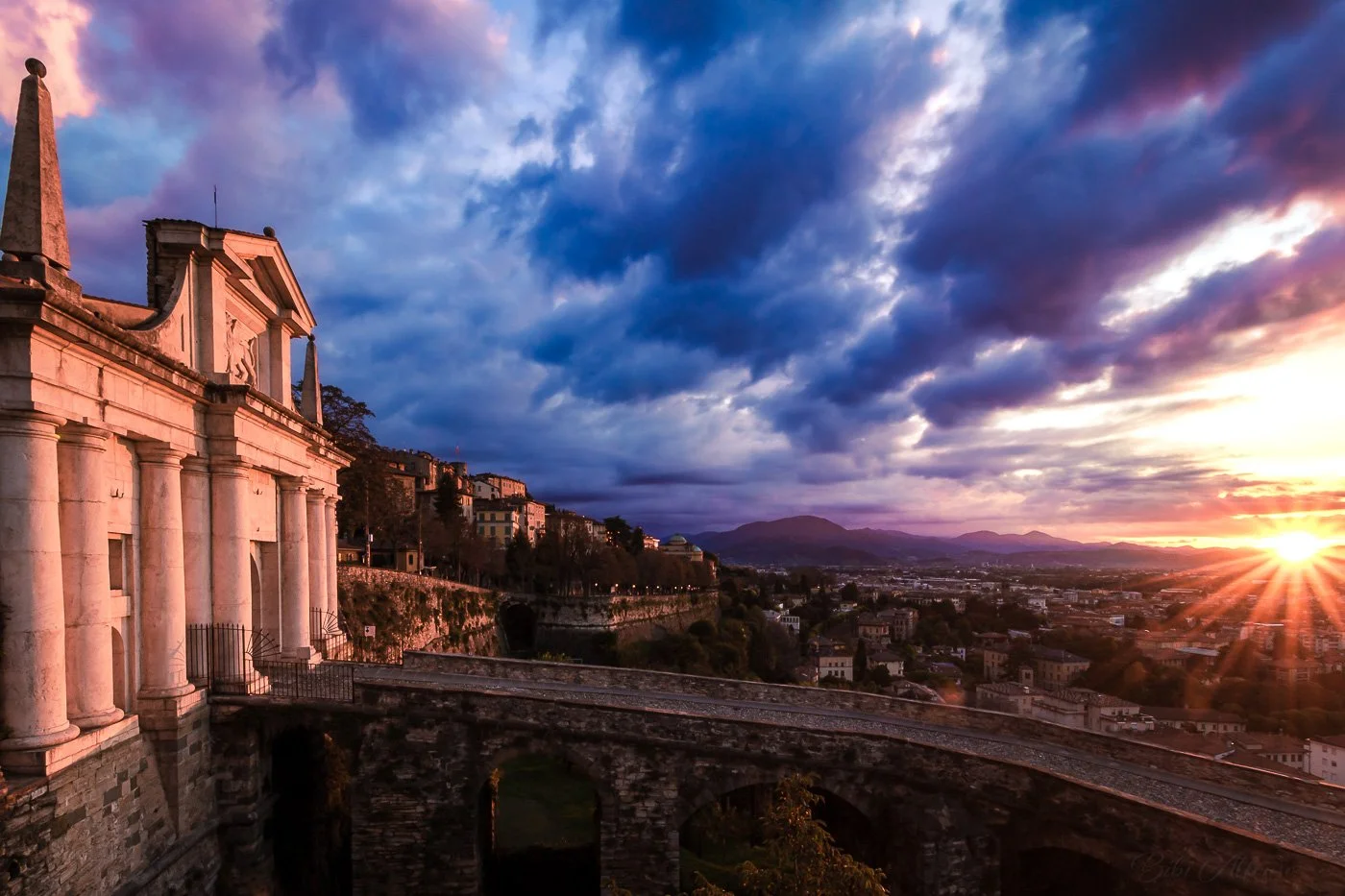 Sunrise illuminating Porta San Giacomo in Bergamo Alta, with warm orange light contrasting the cool blue tones of the early morning sky.
