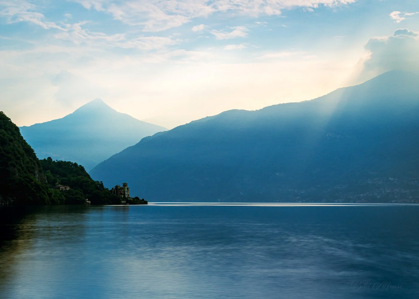 Long exposure photograph of Lake Como, Italy, featuring calm water reflecting lush green hills and mountains on the left under a cloudy sky with soft sunlight.