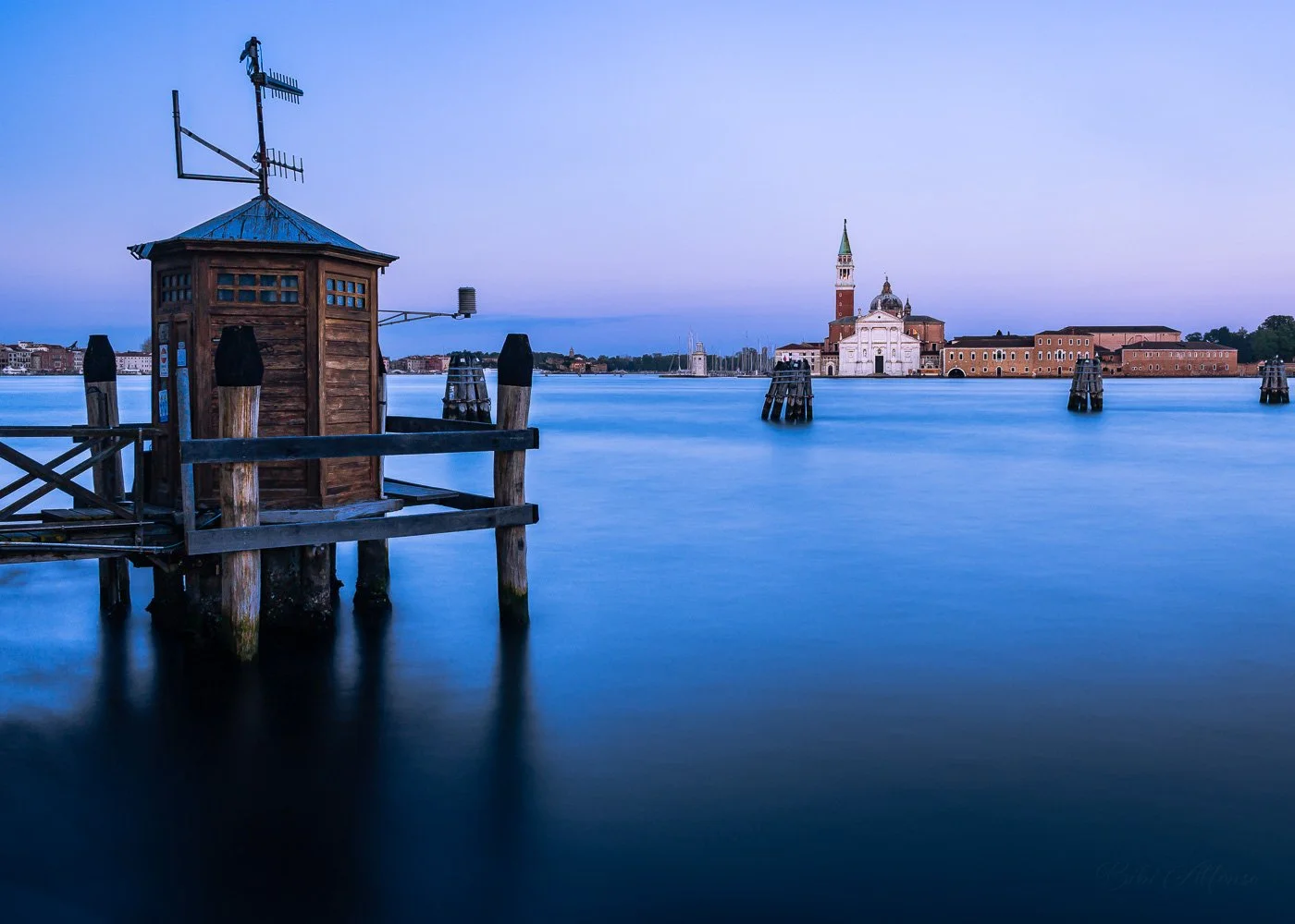 Long exposure afternoon shot from Punta della Dogana, Venice, Italy, featuring a wooden dock with a small hut leading toward the distant cityscape and San Giorgio Maggiore church under a clear sky.
