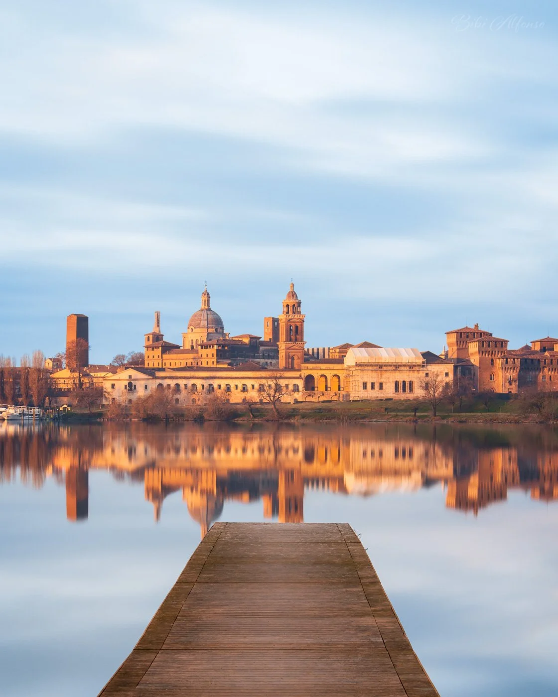 Winter morning skyline of Mantua reflected in calm water, with a wooden pier leading toward historic buildings glowing in warm sunrise light under a soft blue sky.