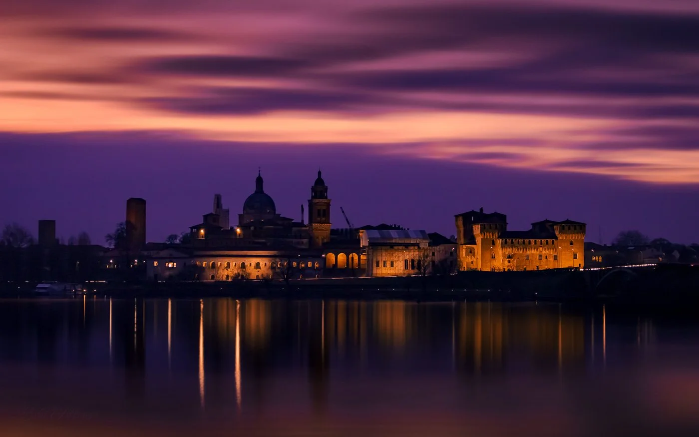 Mantua skyline at blue hour after sunset, long exposure capturing deep blues and purples in the sky, with streetlights illuminating historic buildings and reflections on the calm lake.