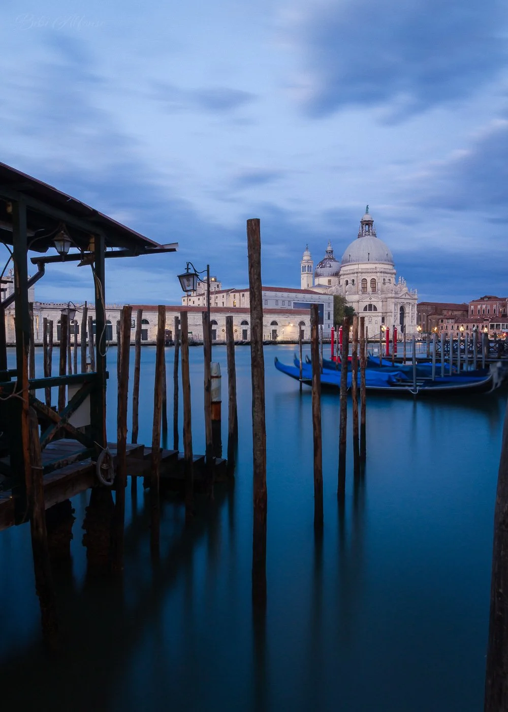 Long exposure blue hour photograph of Venice, Italy, featuring wooden mooring poles, gondolas on the lagoon, and the Basilica di Santa Maria della Salute illuminated in the background.