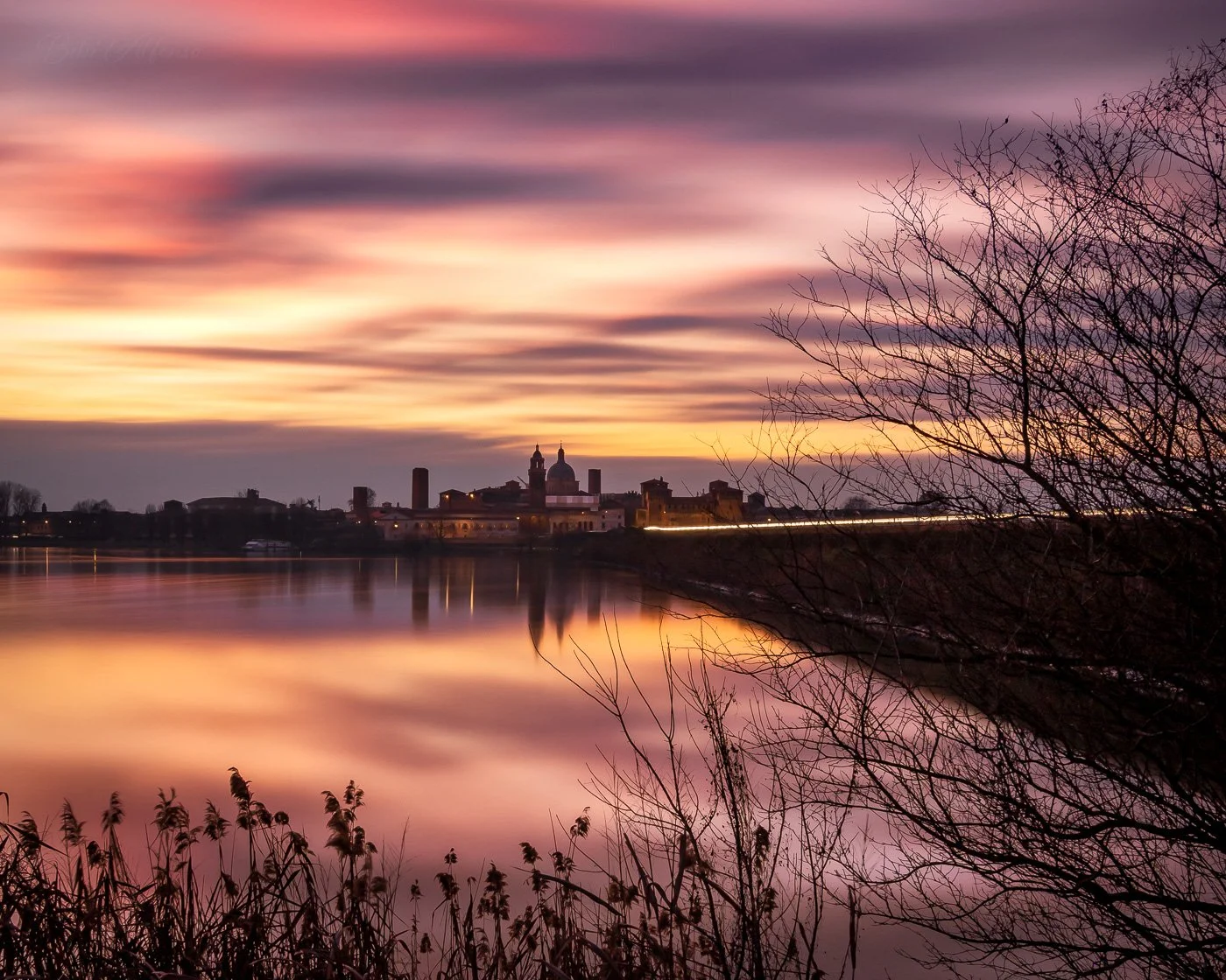 Long exposure photograph of the Mantova skyline at sunset, with pink and gold hues, silky water, blurry skies, and car light trails on the bridge, captured using a neutral density filter.