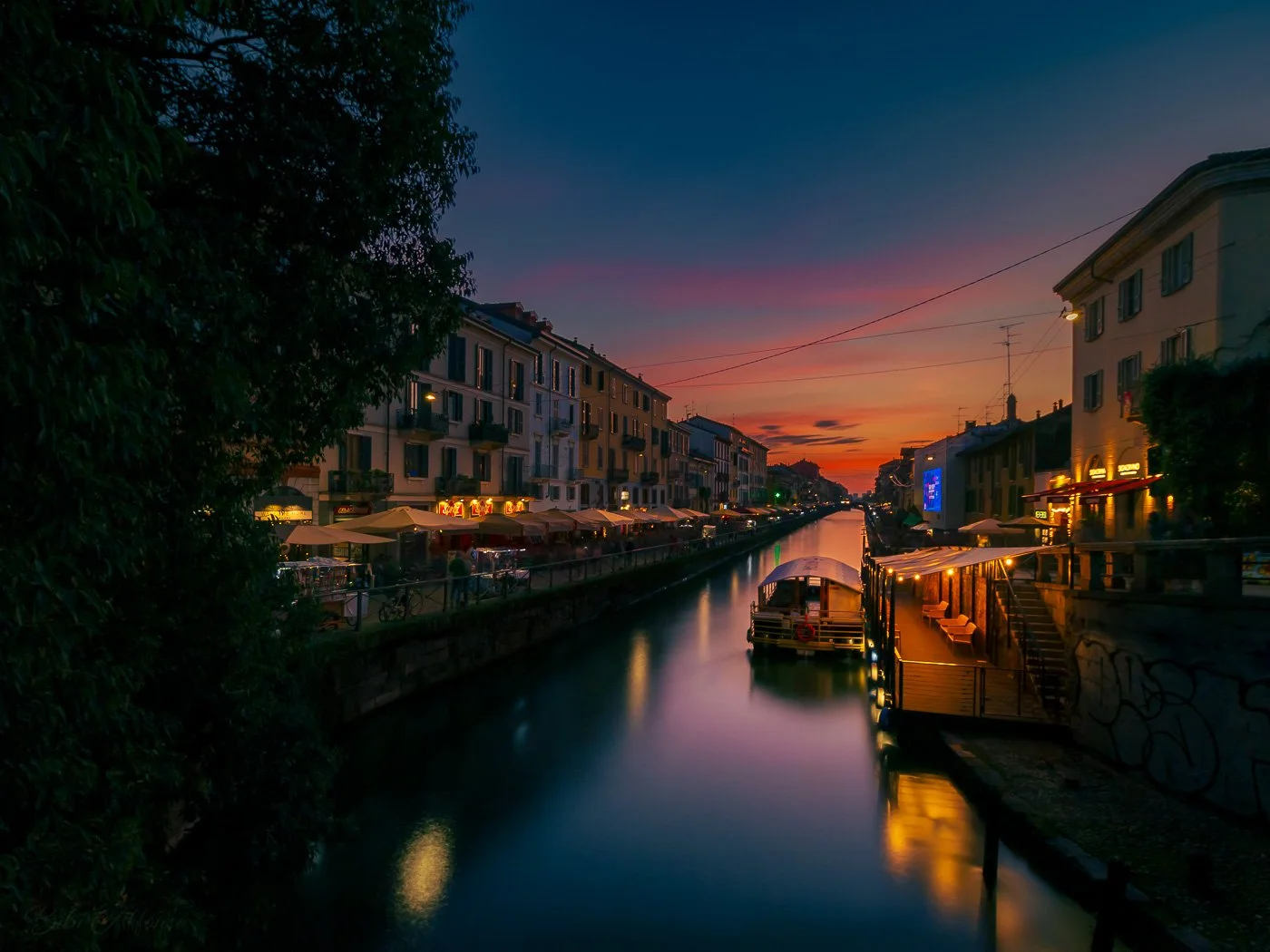 Two minutes long exposure twilight shot of Naviglio Grande canal in Milan, Italy, showing colorful buildings, outdoor cafes with umbrellas, boats, and streetlights glowing under sunset skies.