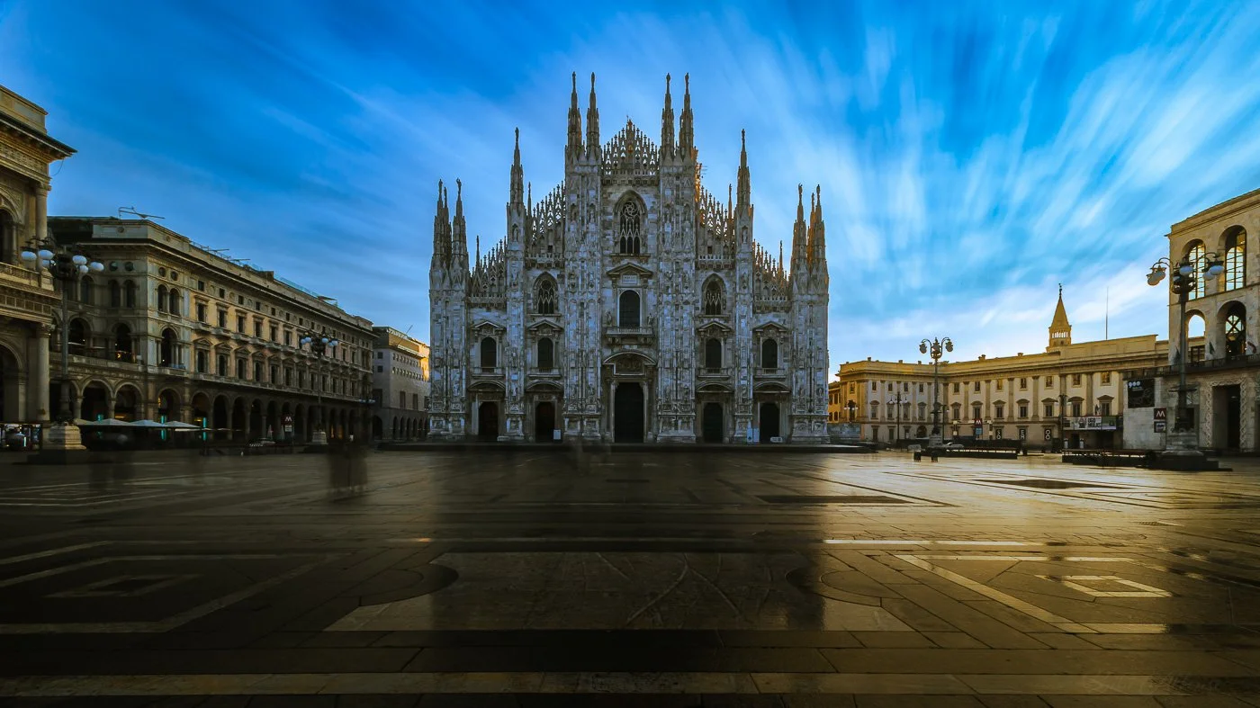 Long exposure photo of Milan’s Duomo with blurred people and streaked clouds, capturing motion around the still cathedral.