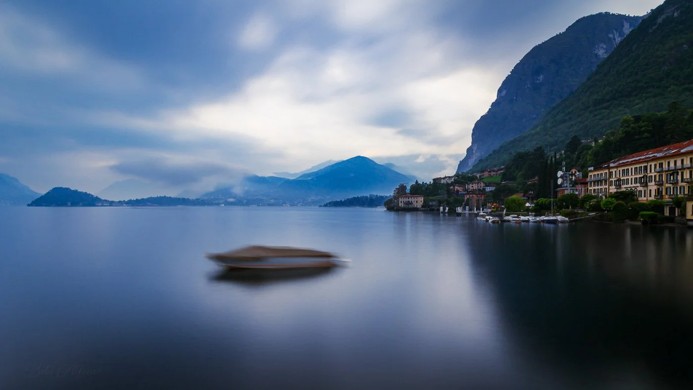 Menaggio on Lake Como beneath a quiet, moody sky, with muted greens and blues reflecting in the lake’s still surface.
